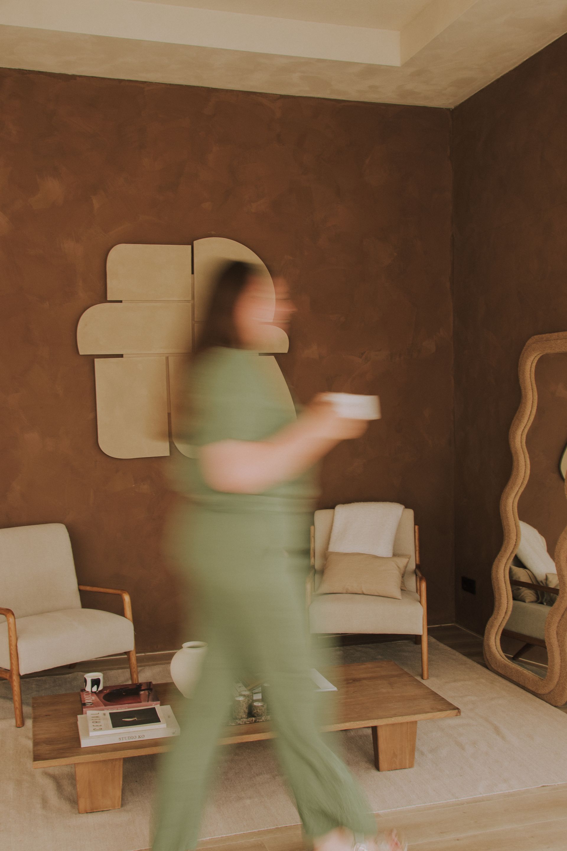 Blurred person in a green outfit walking through a cozy living room carrying a white cup