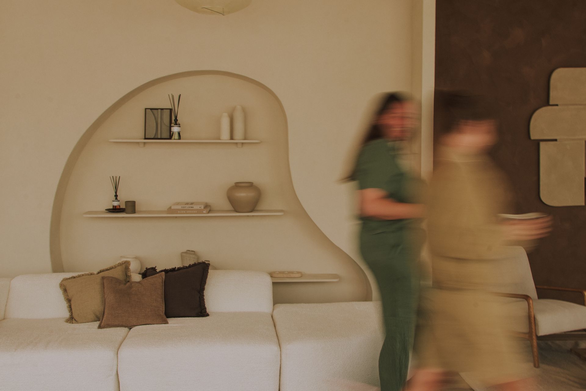 Two blurred people walk past a beige living room with a curved wall shelf and a sofa.