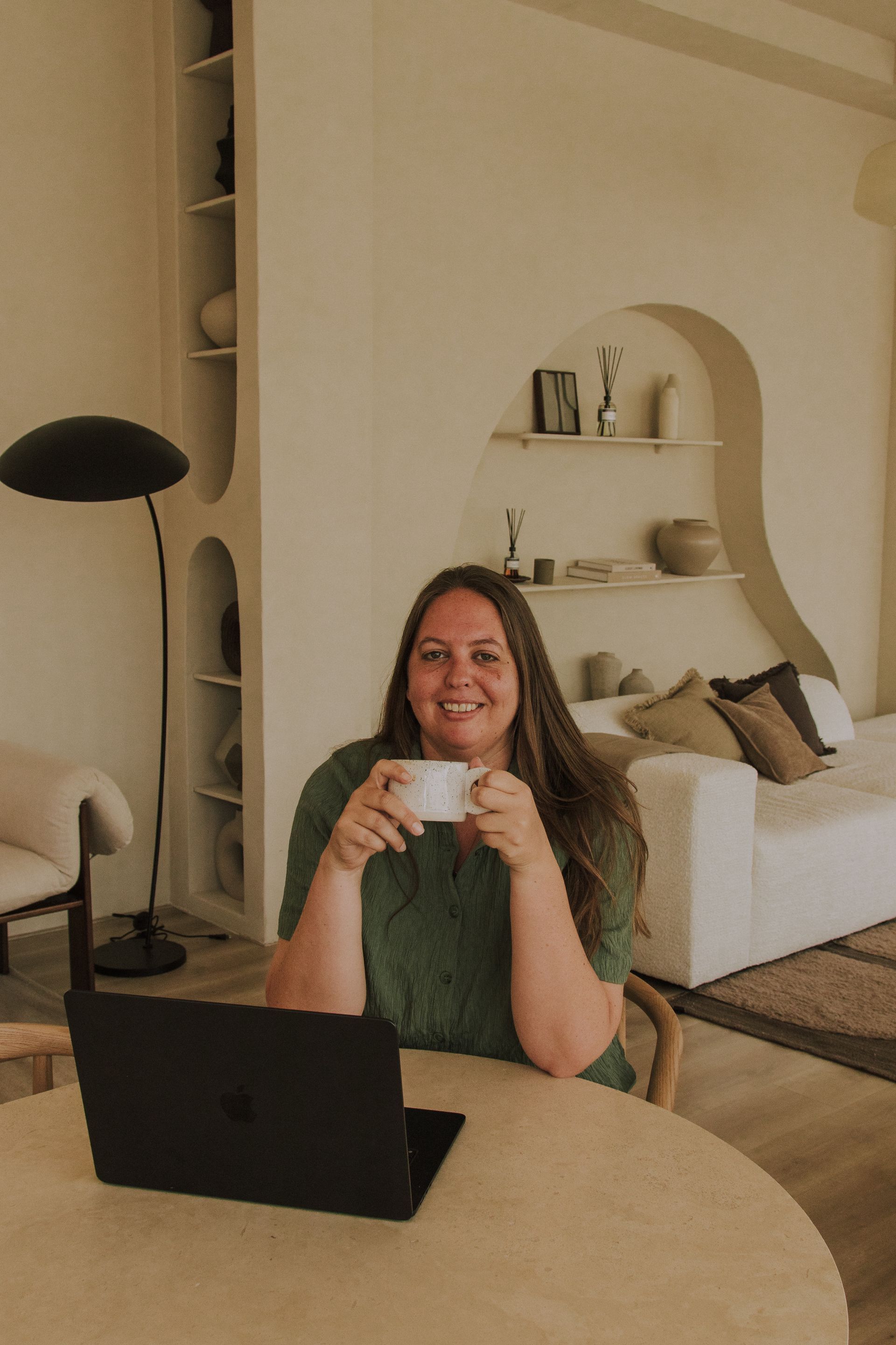 Woman smiling at a round table with a laptop, holding a cup in a cozy cream-colored living room.