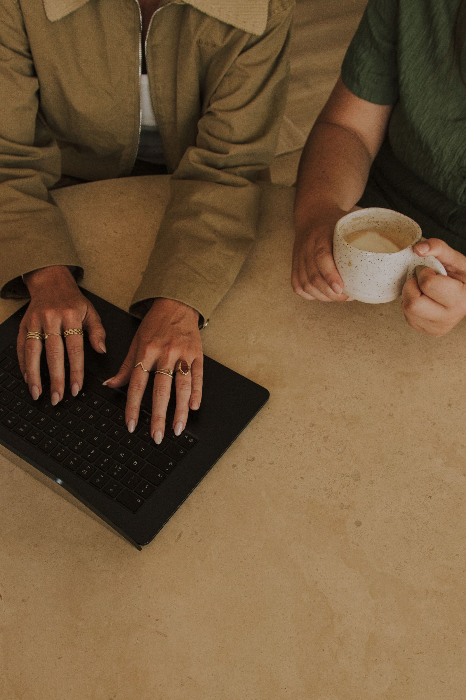 Two people at a table using a laptop, one holding a white mug of coffee.