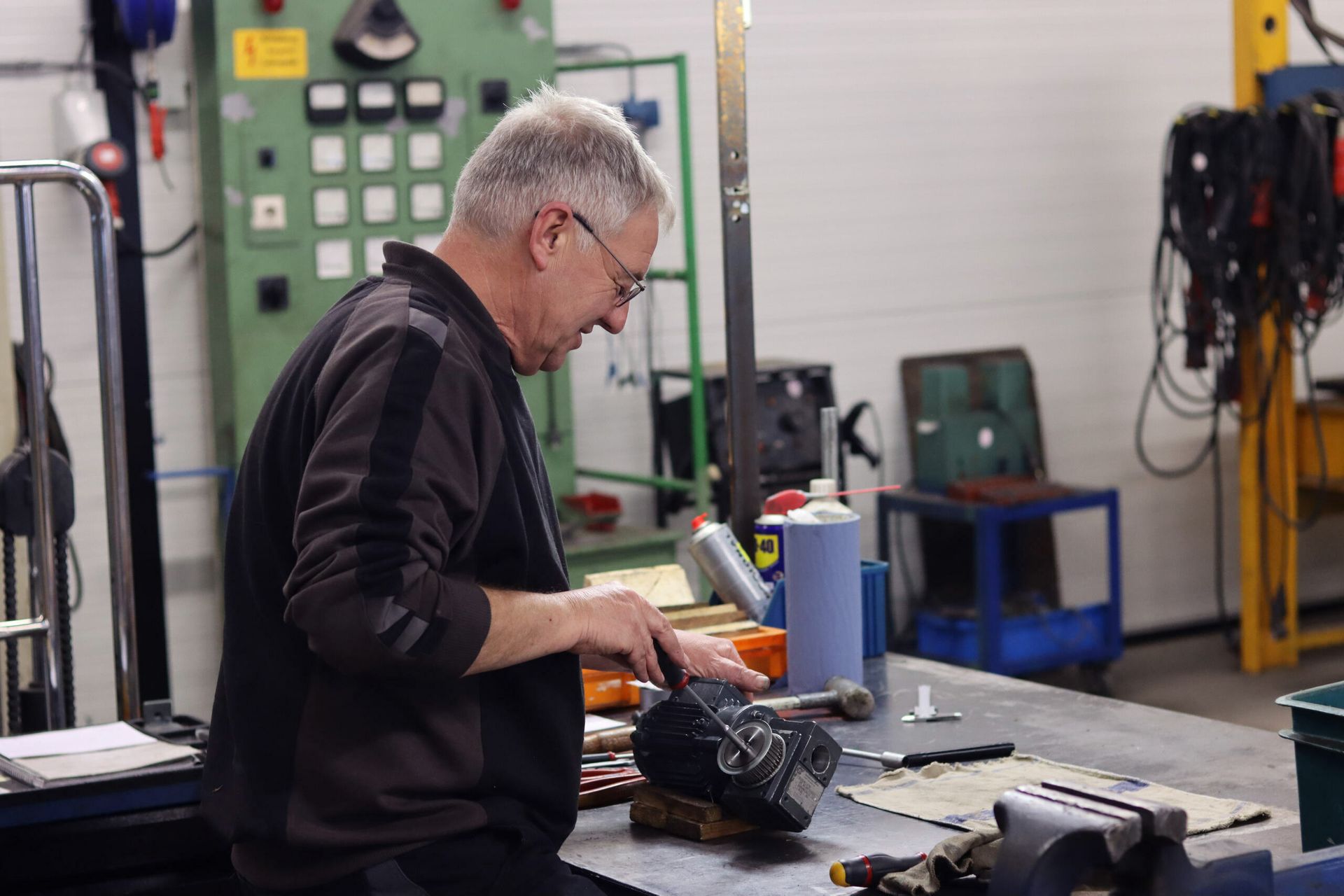 Man working at a workbench in a workshop, using tools near a machine and scattered parts.