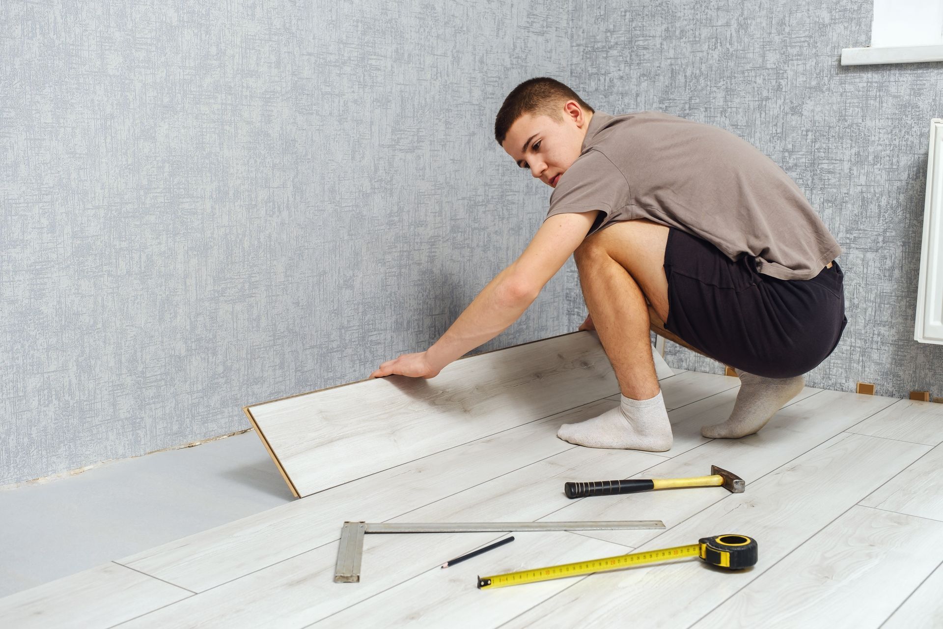 Person installing light-colored floorboards, squatting, tools visible, against a gray wall.