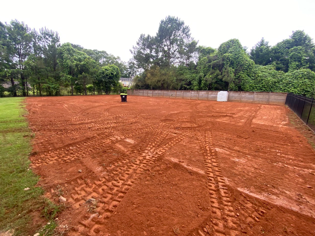 Freshly graded red dirt lot with tire tracks, bordered by grass and trees under a cloudy sky