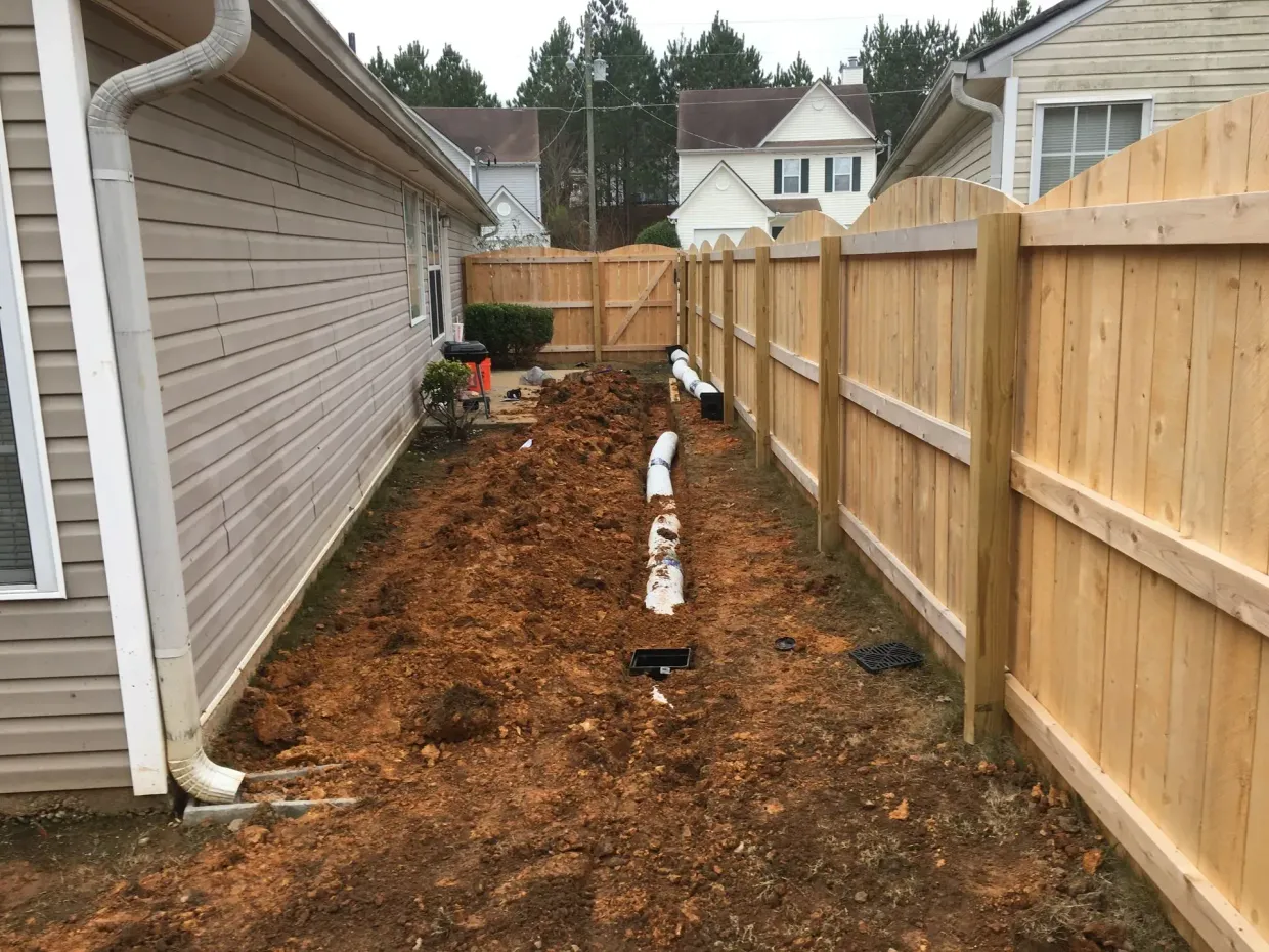 Narrow backyard trench along a house and wooden fence, with exposed white pipes and fresh soil.