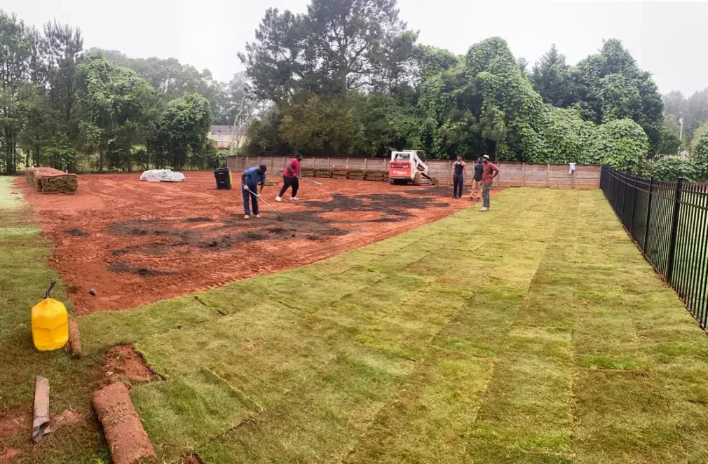 Workers leveling a red dirt field beside a grassy lawn, with trees and a fence in the background.