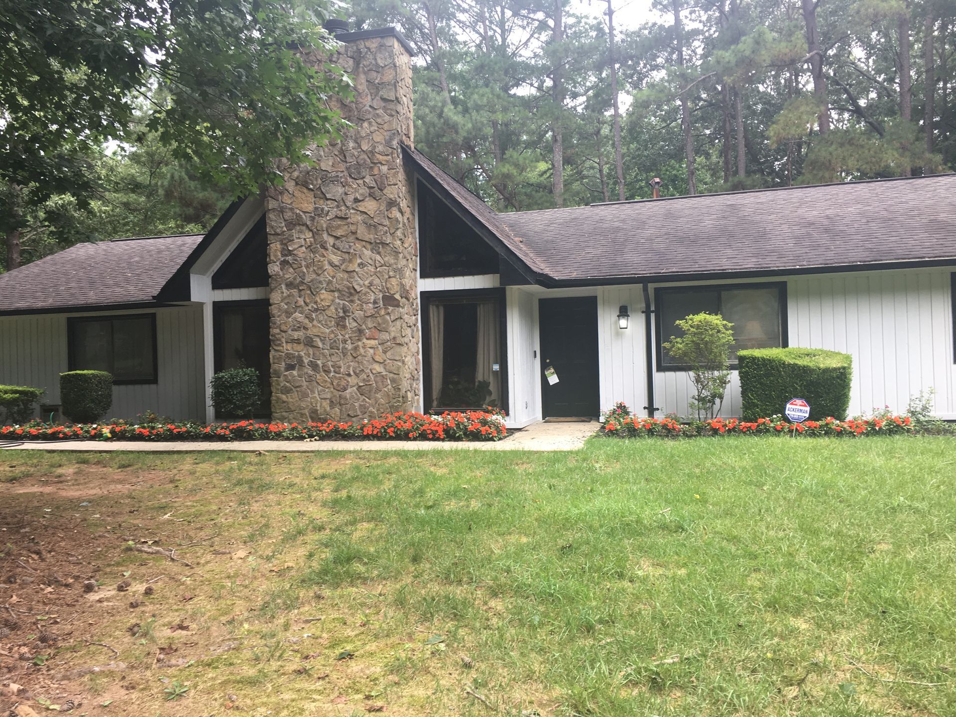 White house with a stone chimney, black trim, and a lawn with flowers and bushes.
