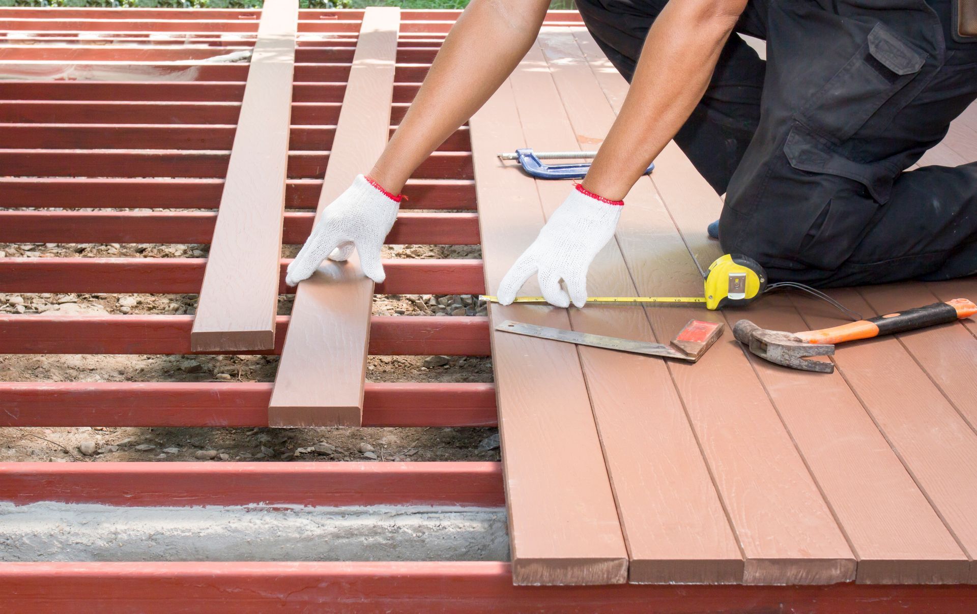 Person installing deck boards on a wooden frame, using a tape measure and gloves.