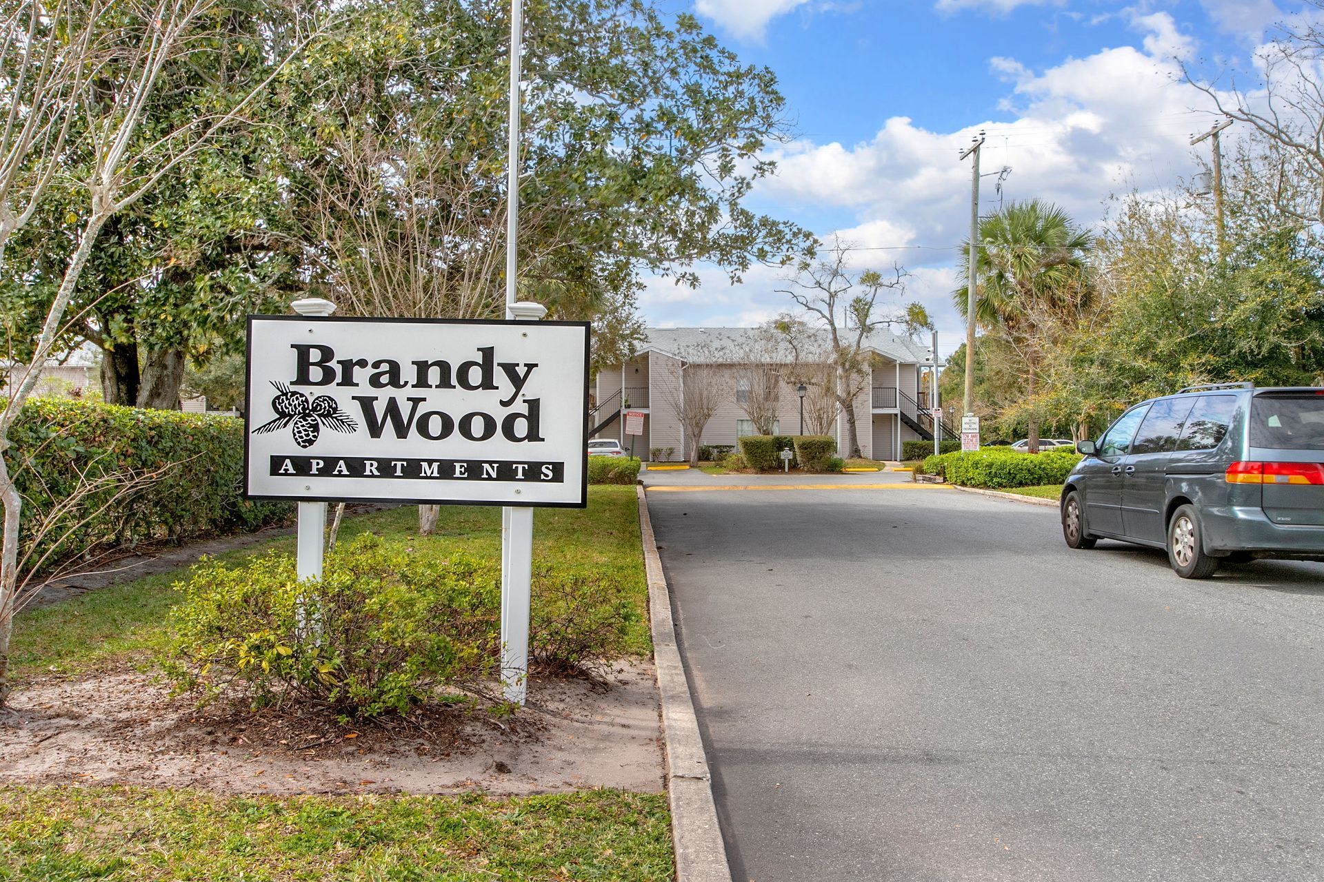 Brandywood Apartments entrance sign beside a road with trees and a parked gray car