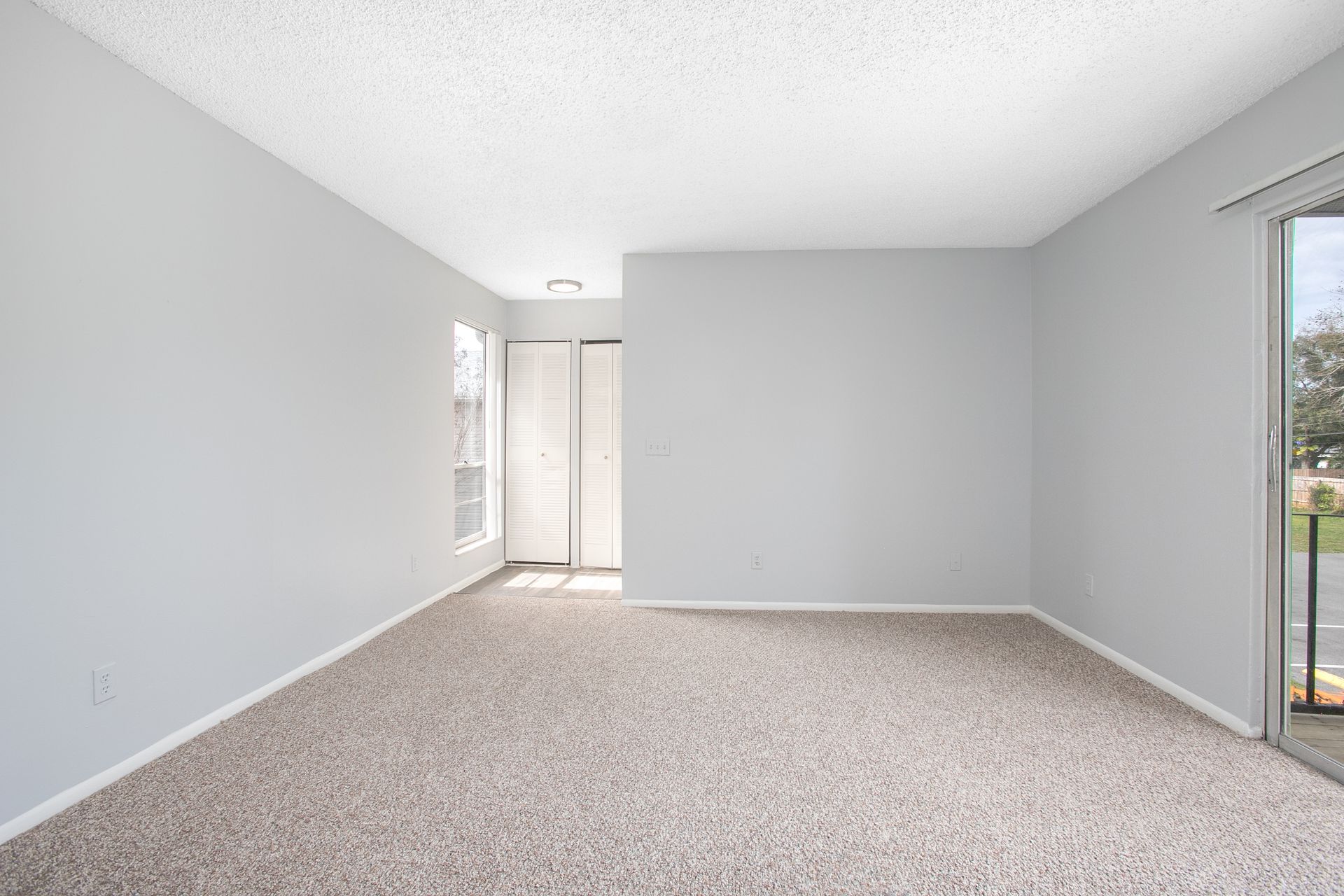 Empty carpeted living room with light gray walls, sliding glass door, and small windows.