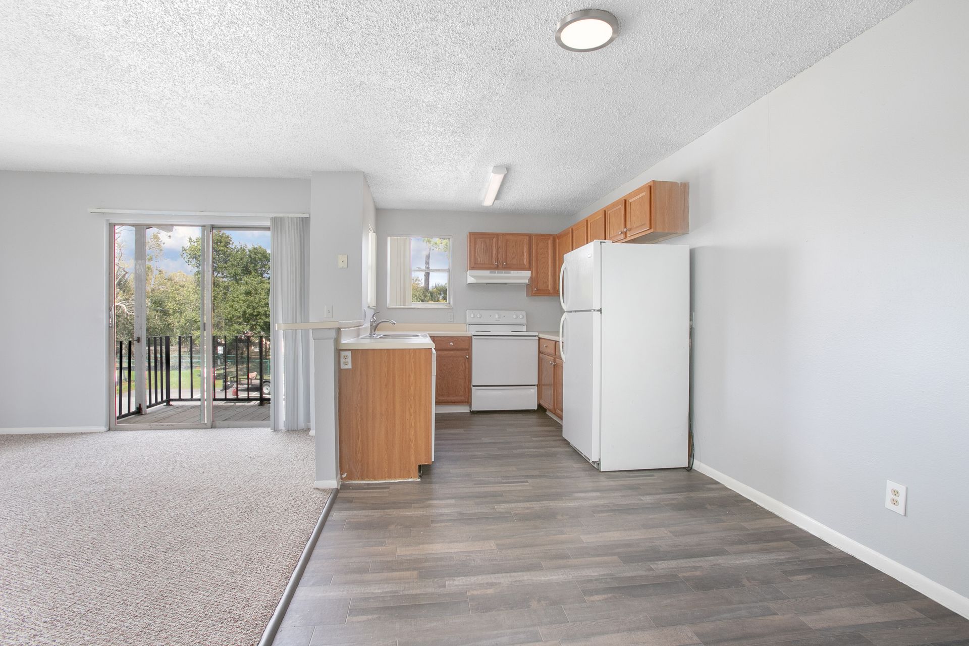 Bright open kitchen and living room with white appliances, wood cabinets, gray floors, and sliding glass doors to a balcony
