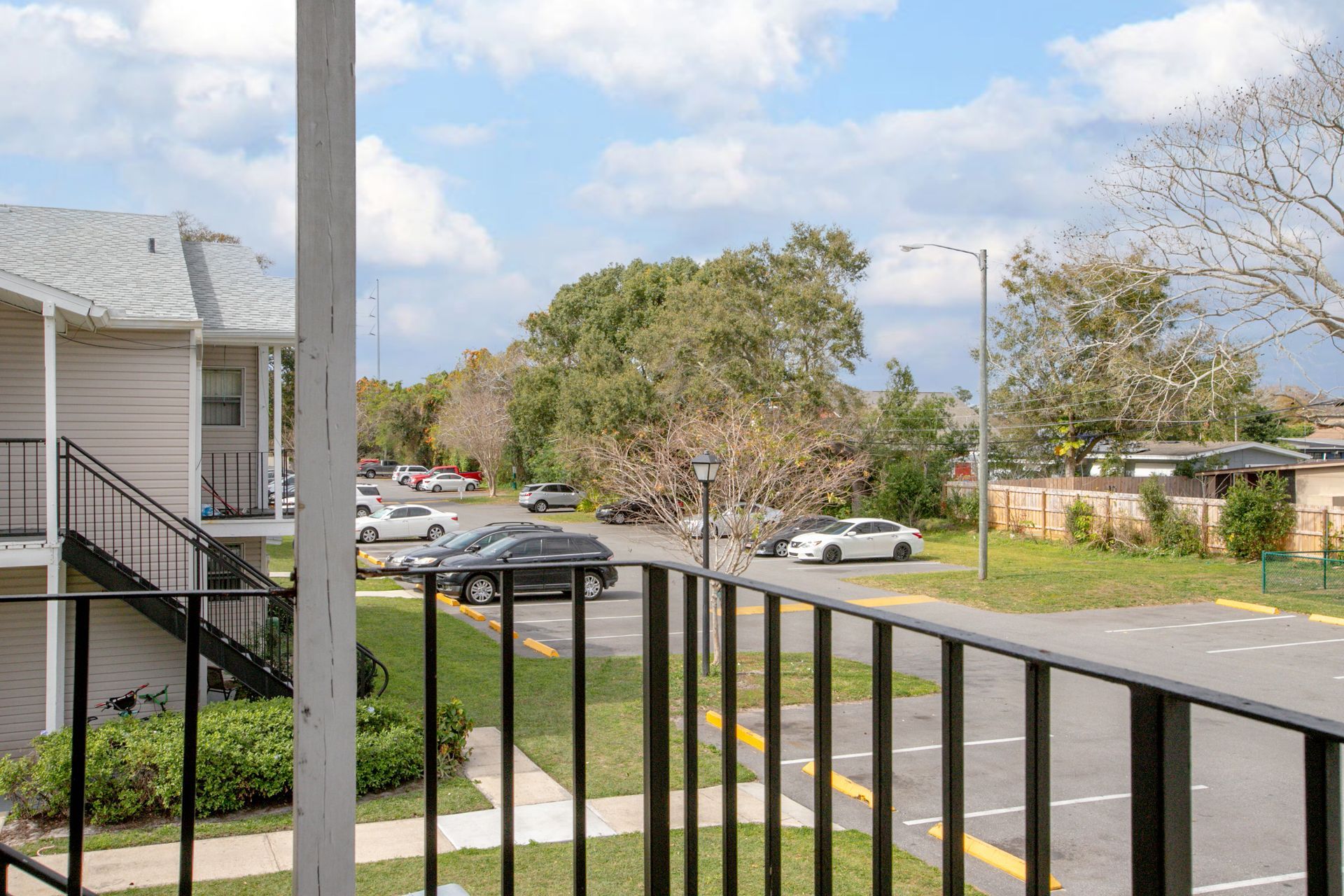 Apartment balcony view of a quiet street, parked cars, trees, and a partly cloudy sky