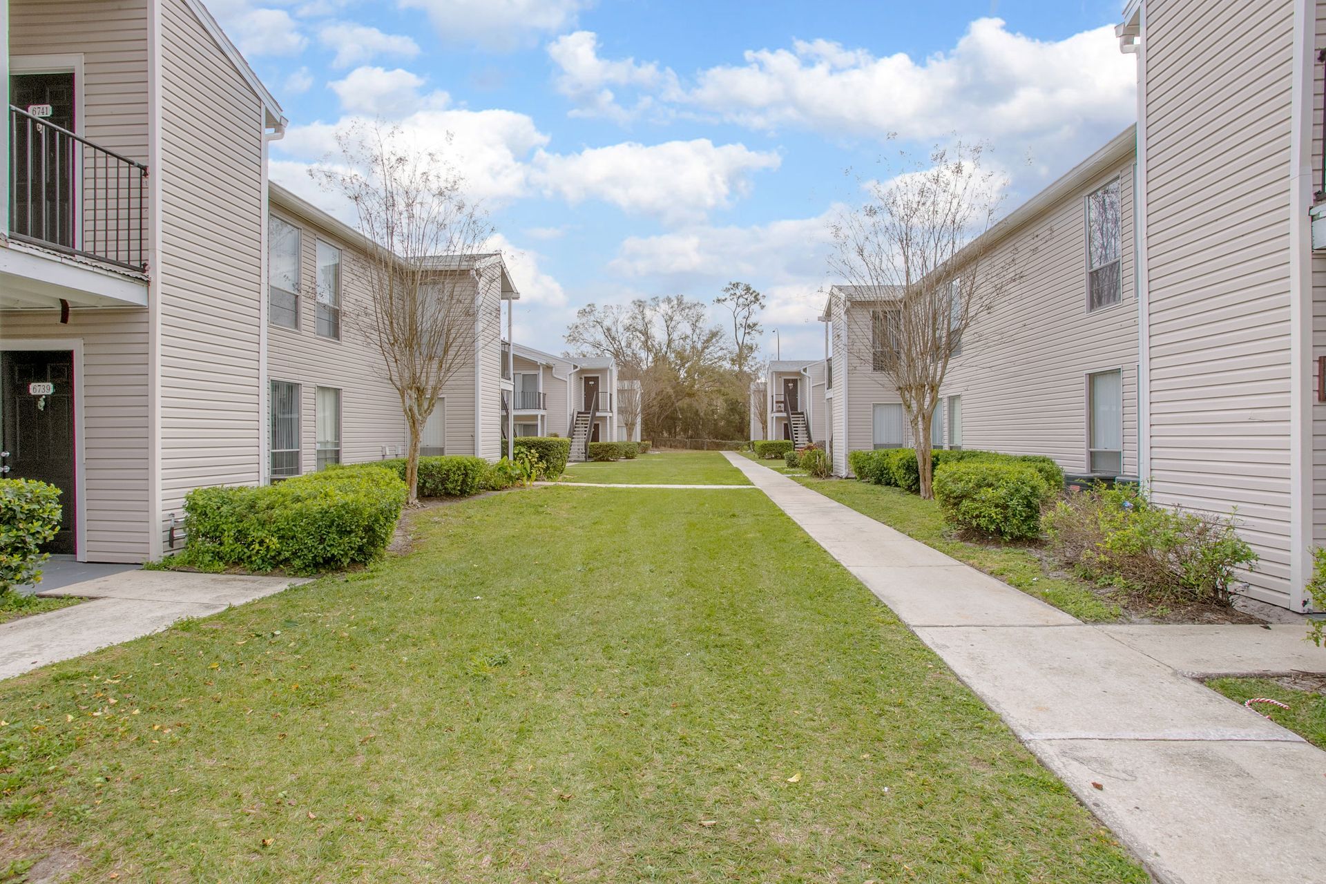 Apartment courtyard with a grassy path between beige buildings and sidewalks under a partly cloudy sky