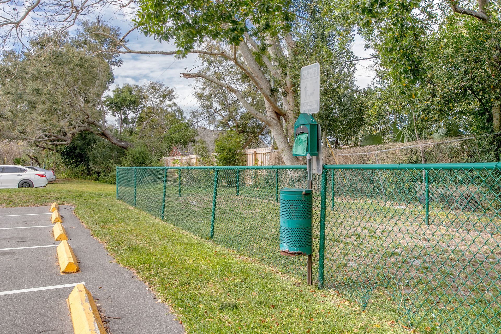 Green chain-link fence along a sidewalk beside a parking lot, with trees and a utility pole.