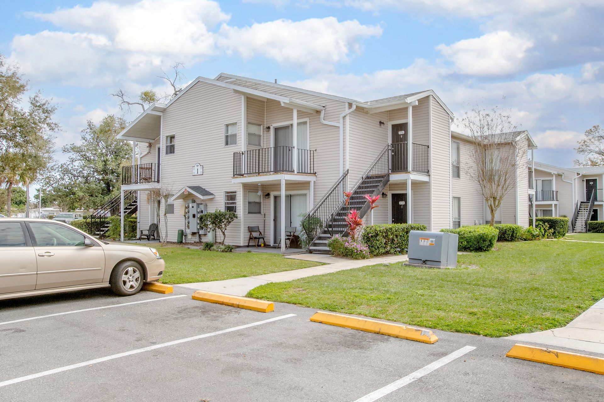Apartment complex with beige siding, exterior stairs, parked car, and grassy lawn under a cloudy sky
