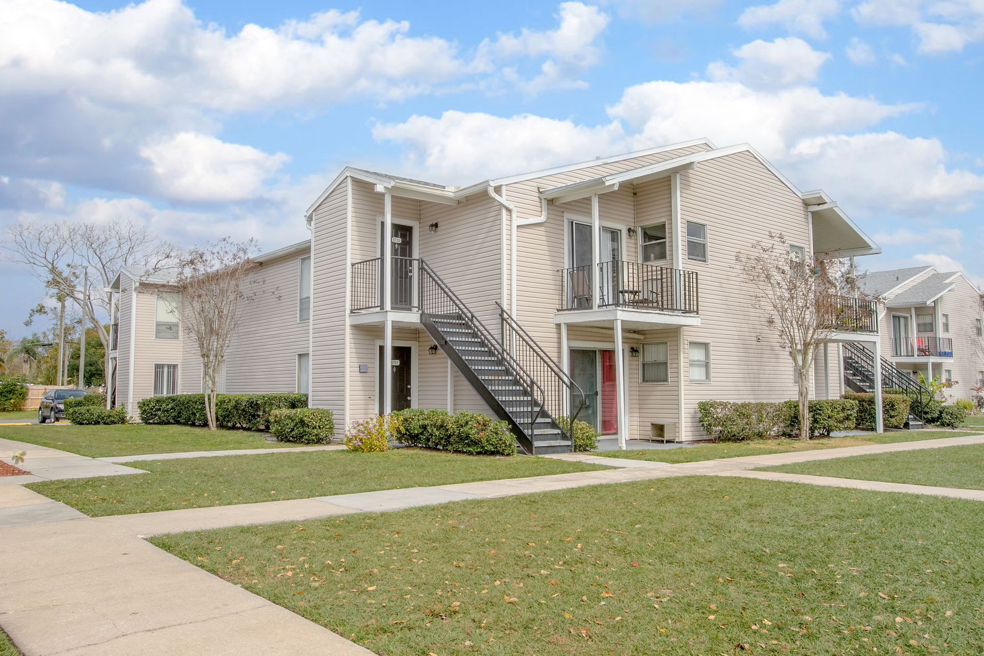 Apartment complex with beige siding, exterior staircases, and a grassy courtyard under a partly cloudy sky