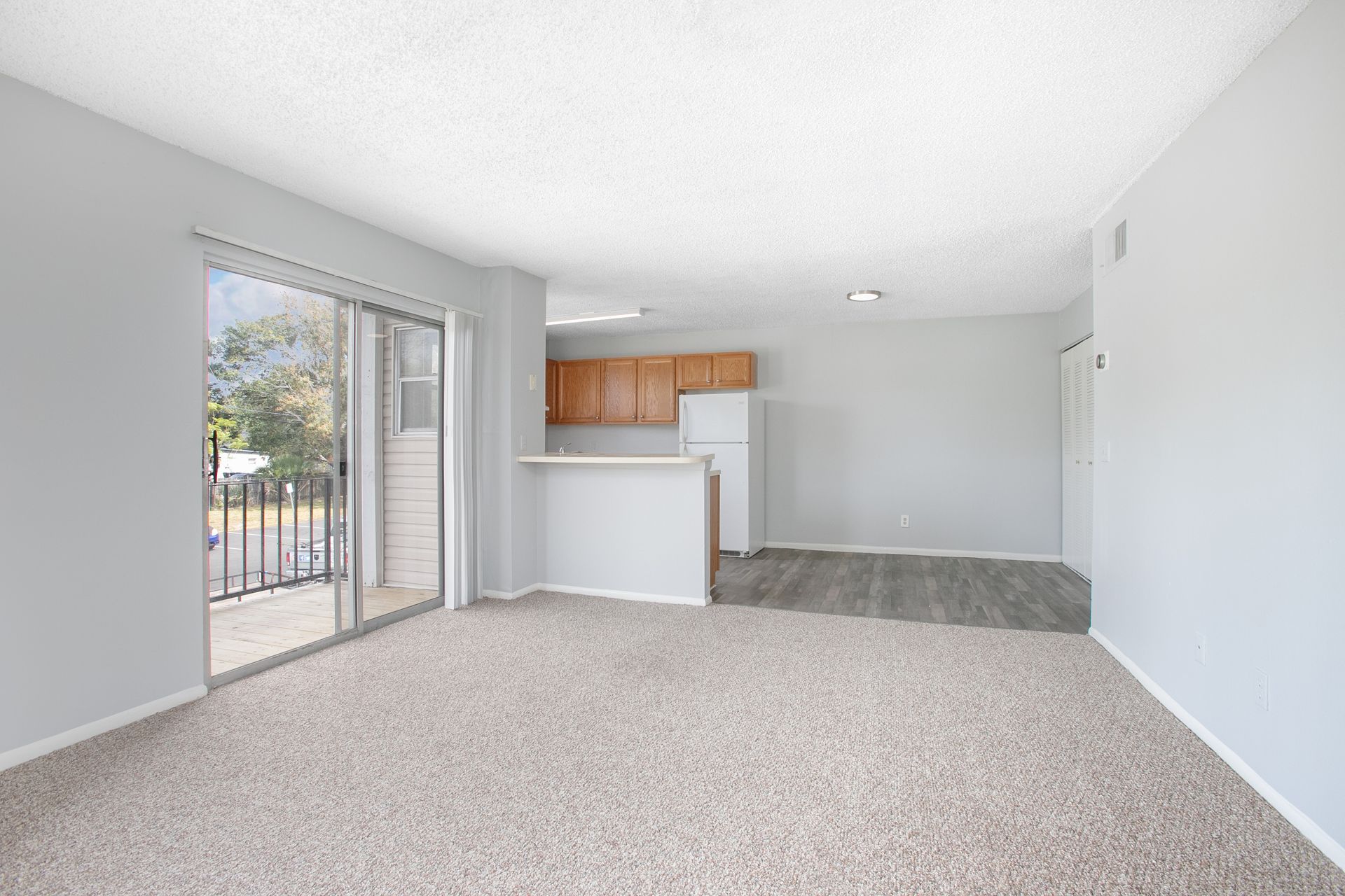 Empty living room with carpet, white walls, and a small kitchen area with wood cabinets.