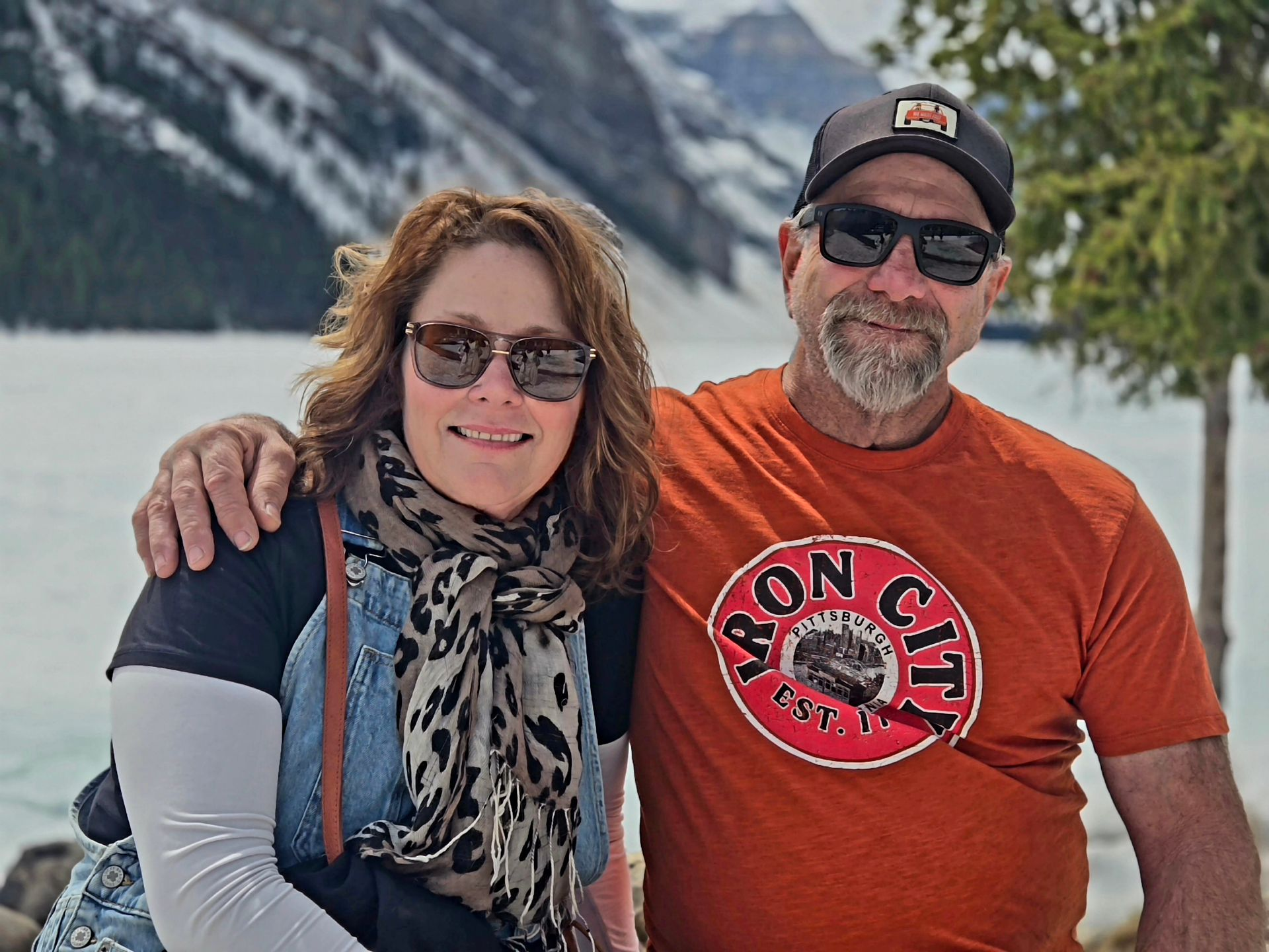 Two smiling people posing in front of a mountain lake, wearing sunglasses and casual clothes.