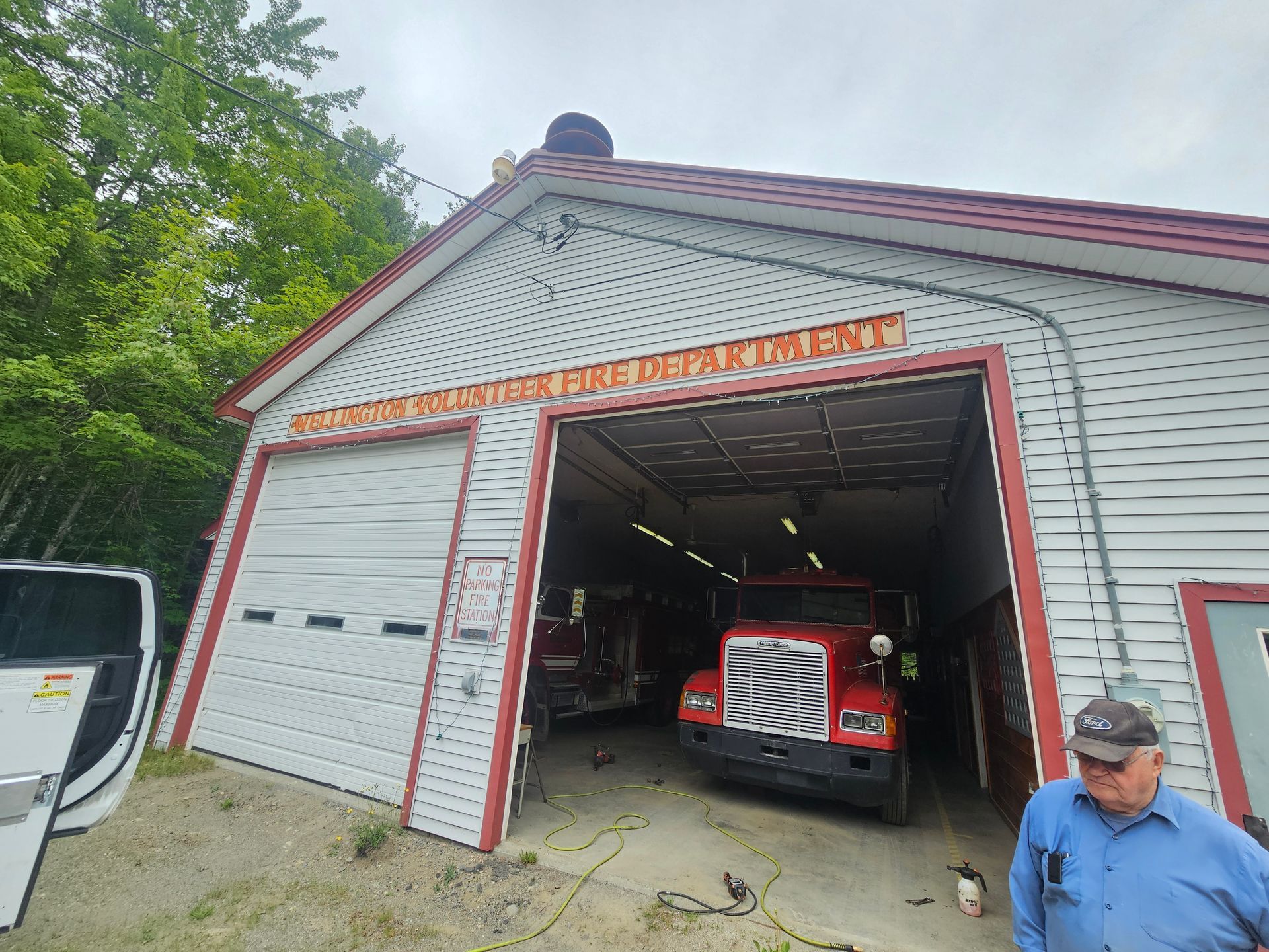 A man is standing in front of a fire station with two fire trucks parked in the garage.