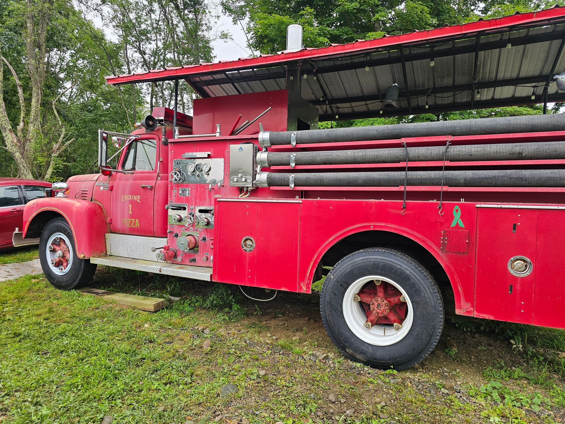 An old red fire truck is parked in the grass under a roof.
