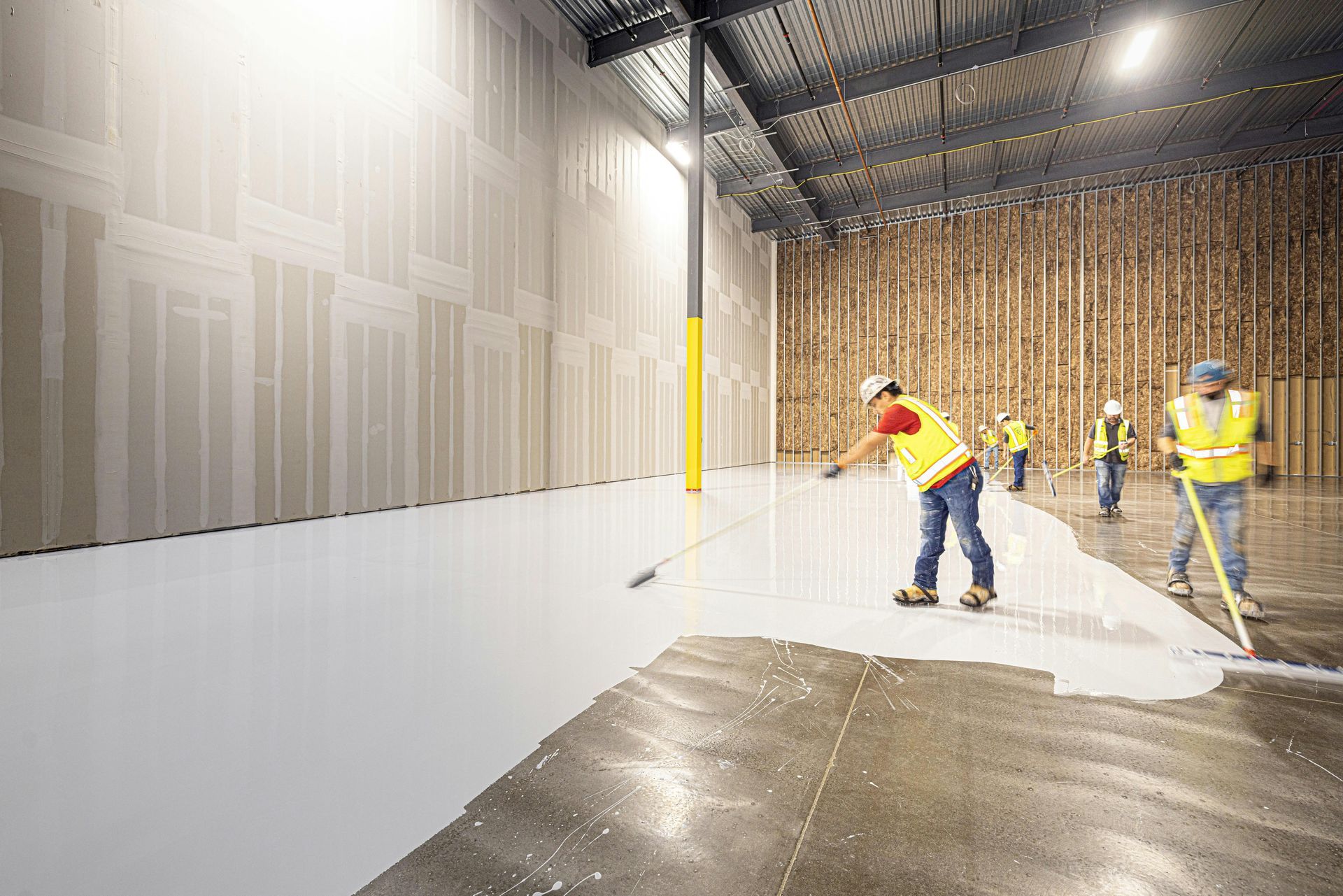 Construction workers troweling white floor coating inside a warehouse with a wet concrete area in foreground