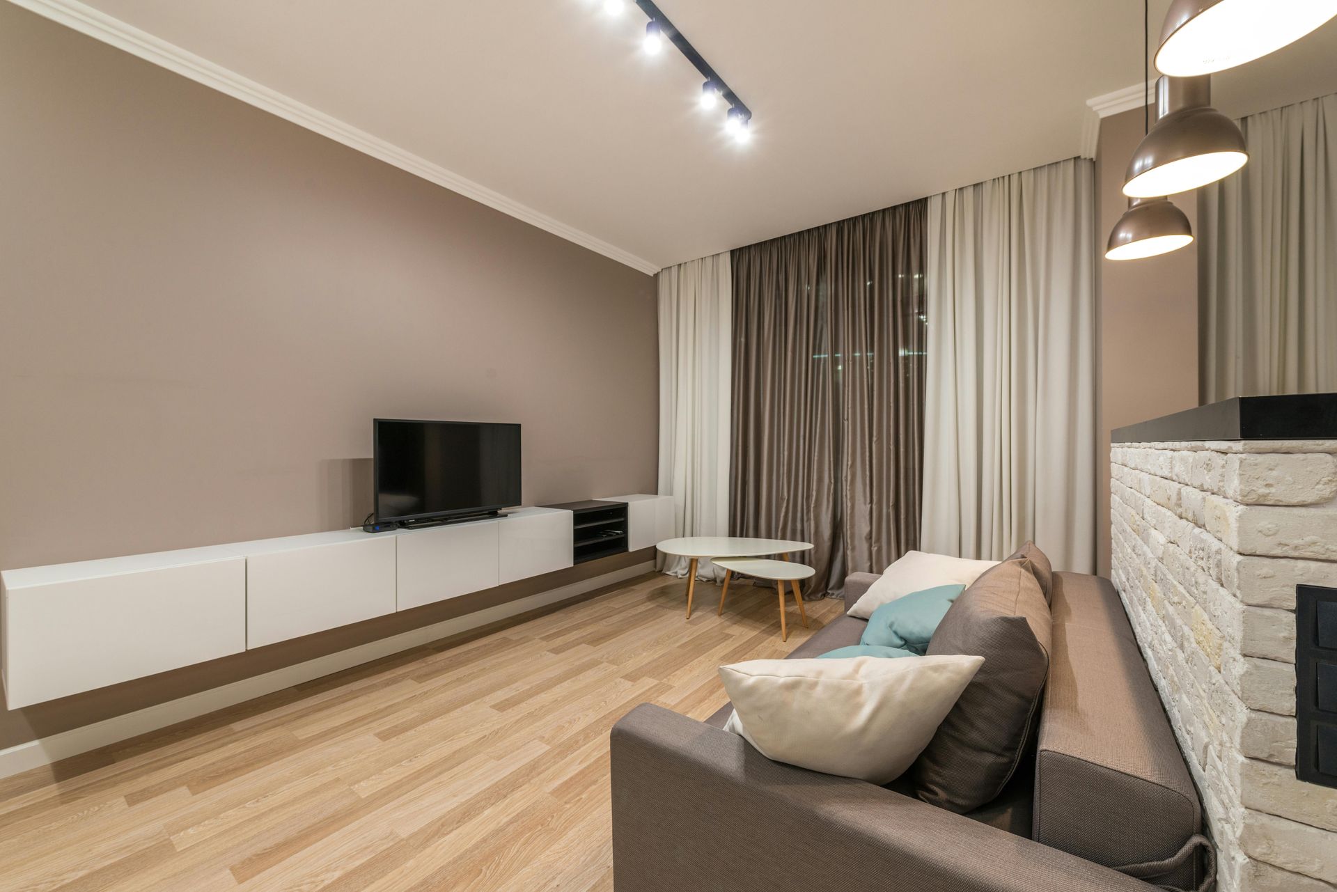 Modern living room with gray sofa, wall-mounted TV, wood floor, and tall windows with curtains