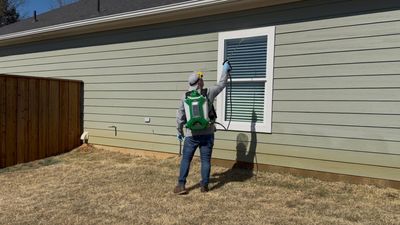Worker in green vest sprays paint on a house window frame beside a wooden fence.
