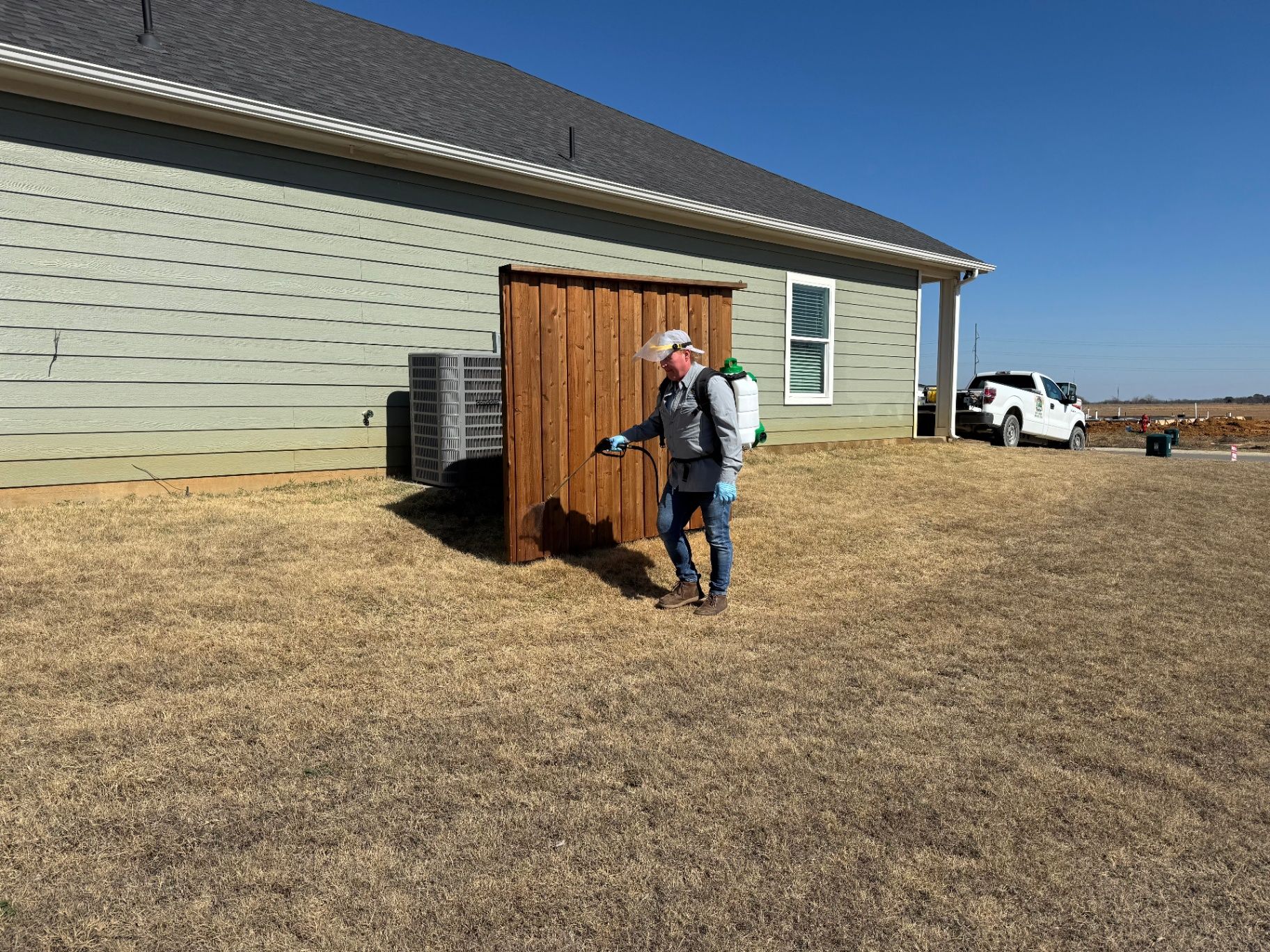A technician uses a handheld sprayer to apply a coating to a wooden privacy screen next to a house on a sunny day.