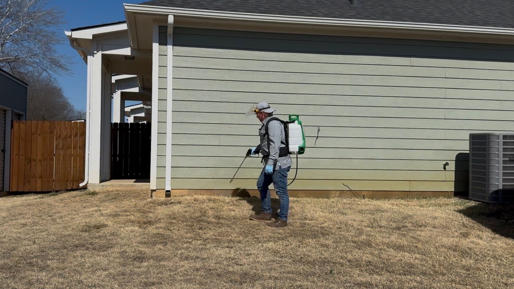 Worker spraying yard near a beige house with a backpack sprayer