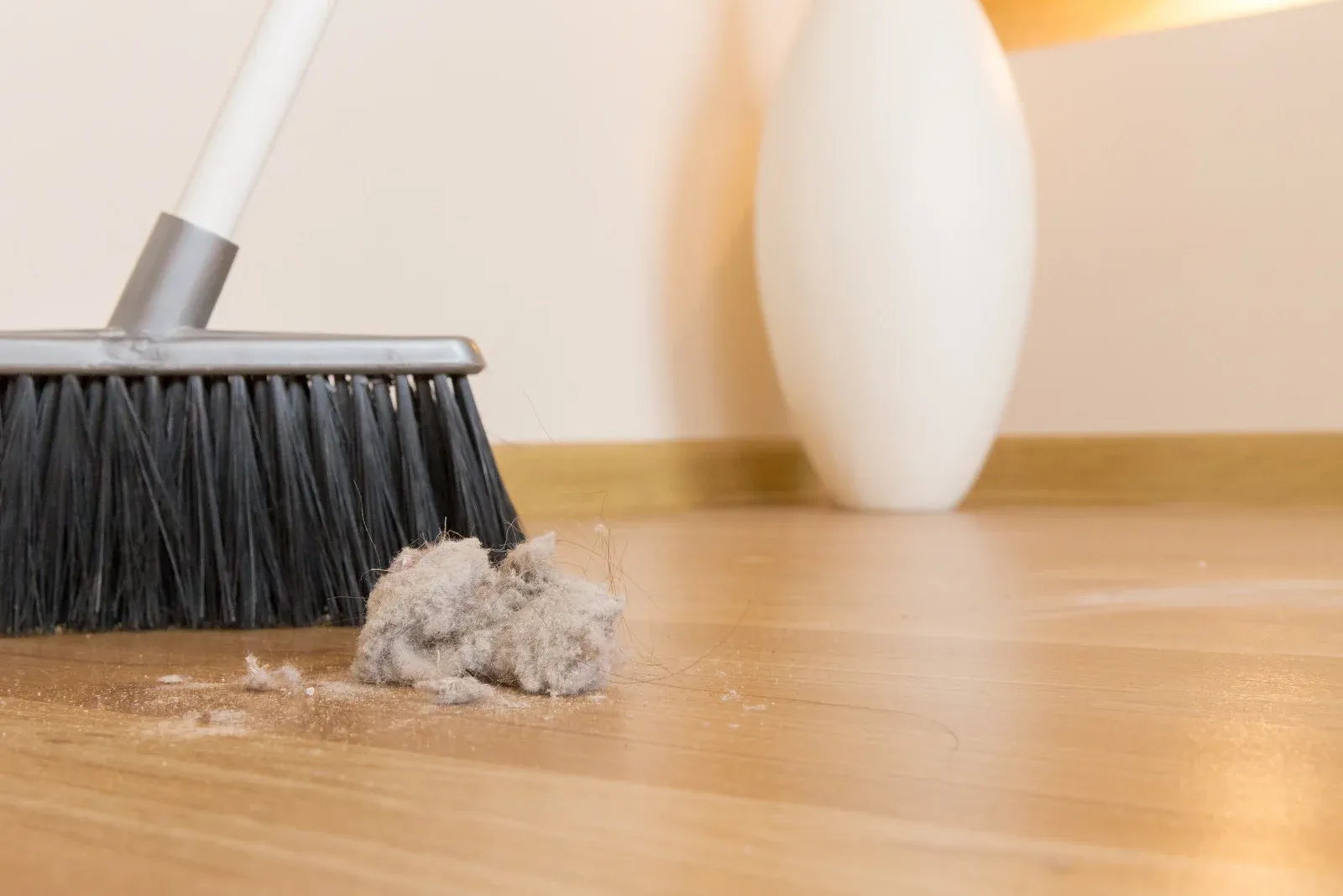 Broom sweeping dust and debris from a wooden floor beside a white vase