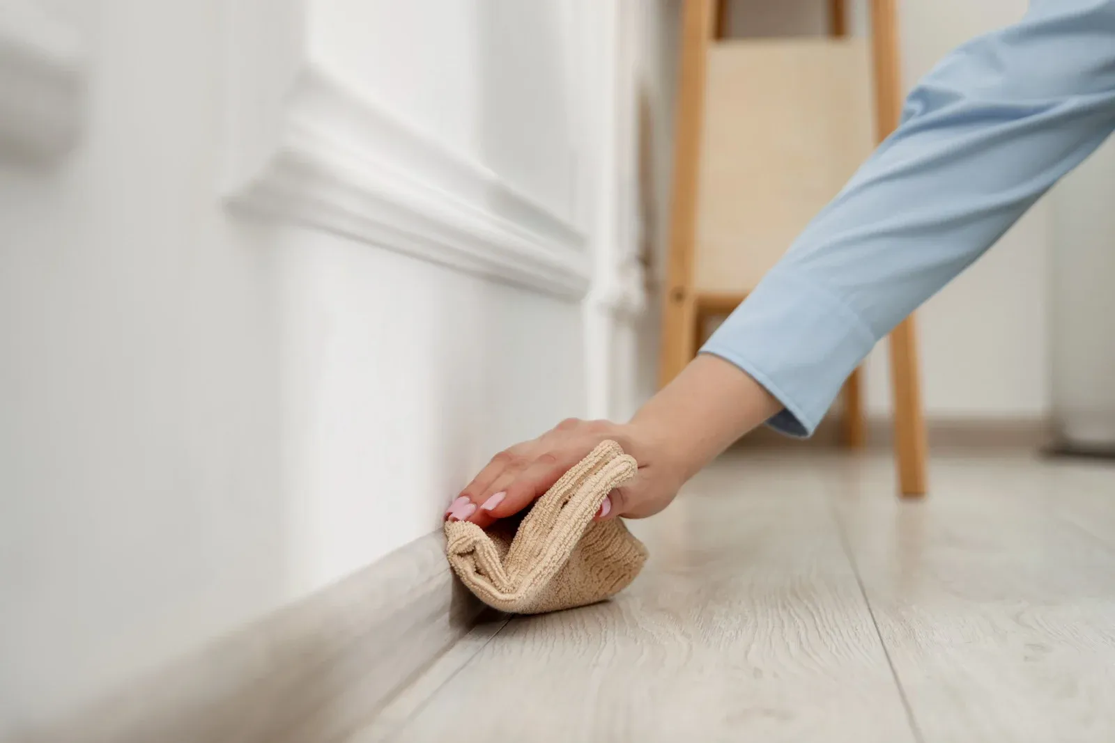 Hand wiping a wooden floor with a cloth near a white wall.