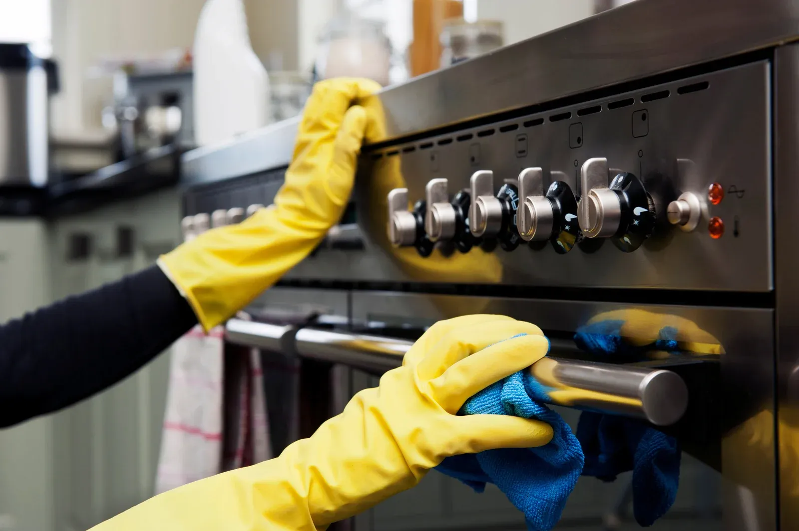 Person cleaning a stainless steel oven with yellow gloves and a blue cloth