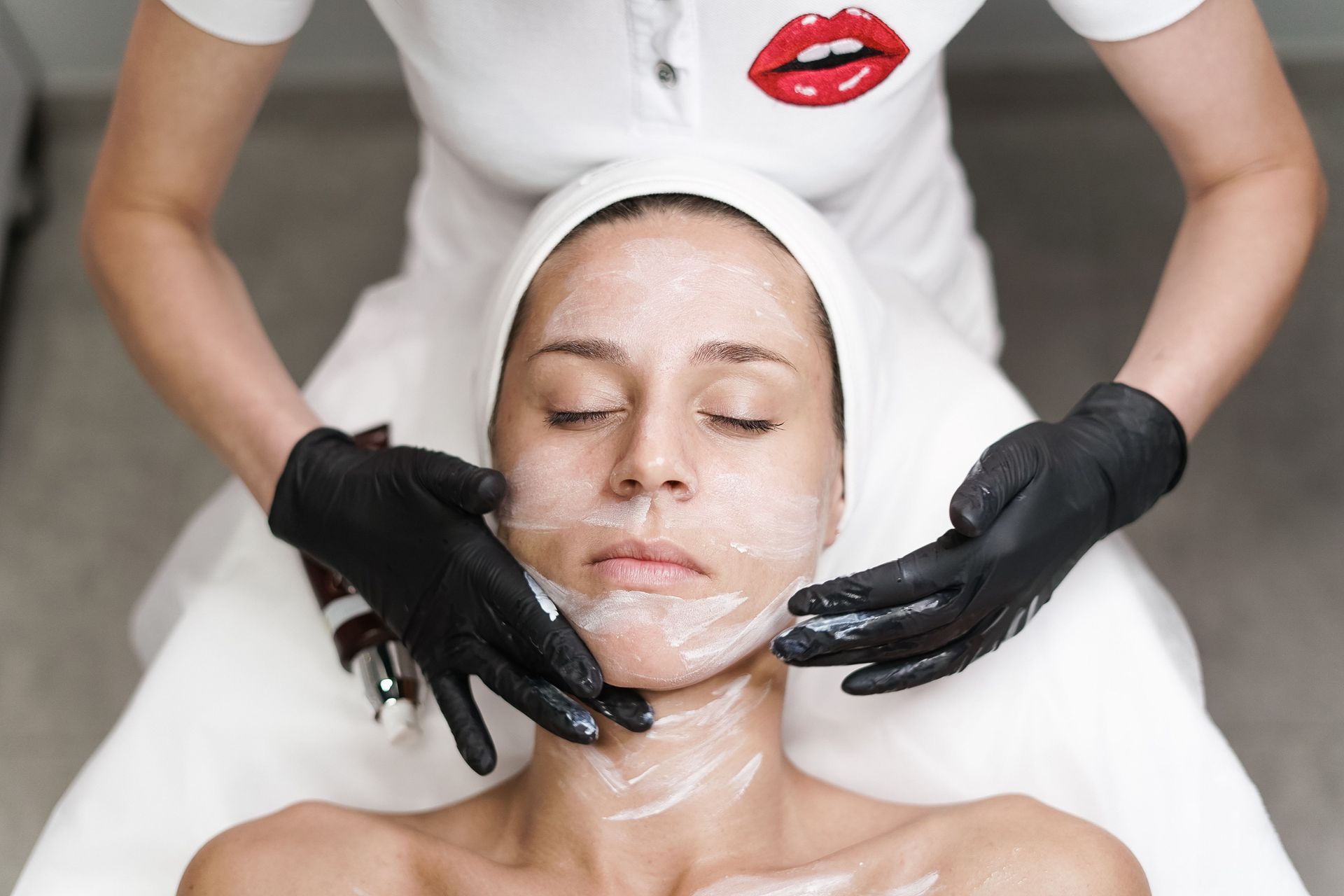 Tratamiento facial con manos enguantadas de negro aplicando crema en el rostro de una mujer en un entorno de spa.