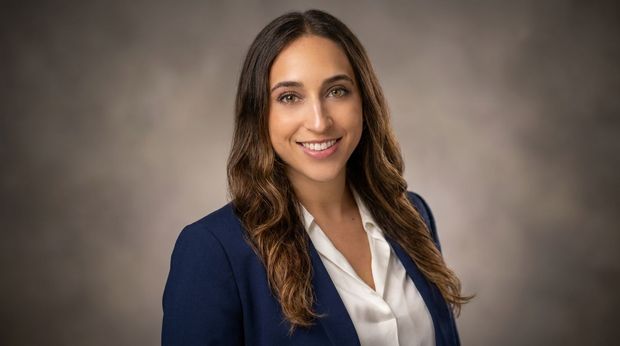 Professional headshot of a smiling woman in a navy blazer and white blouse against a gray backdrop