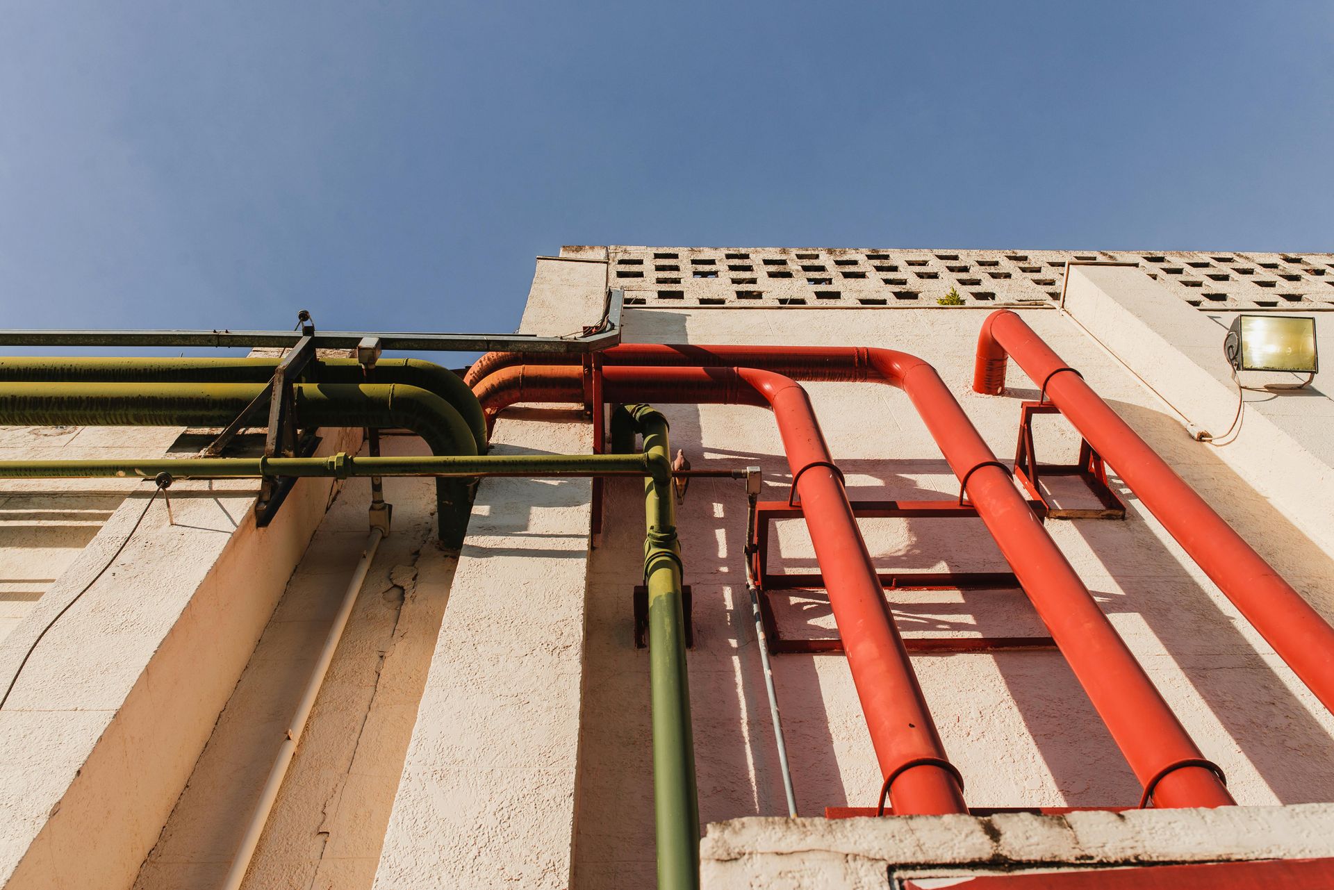 Red and green pipes running up a beige building against a blue sky