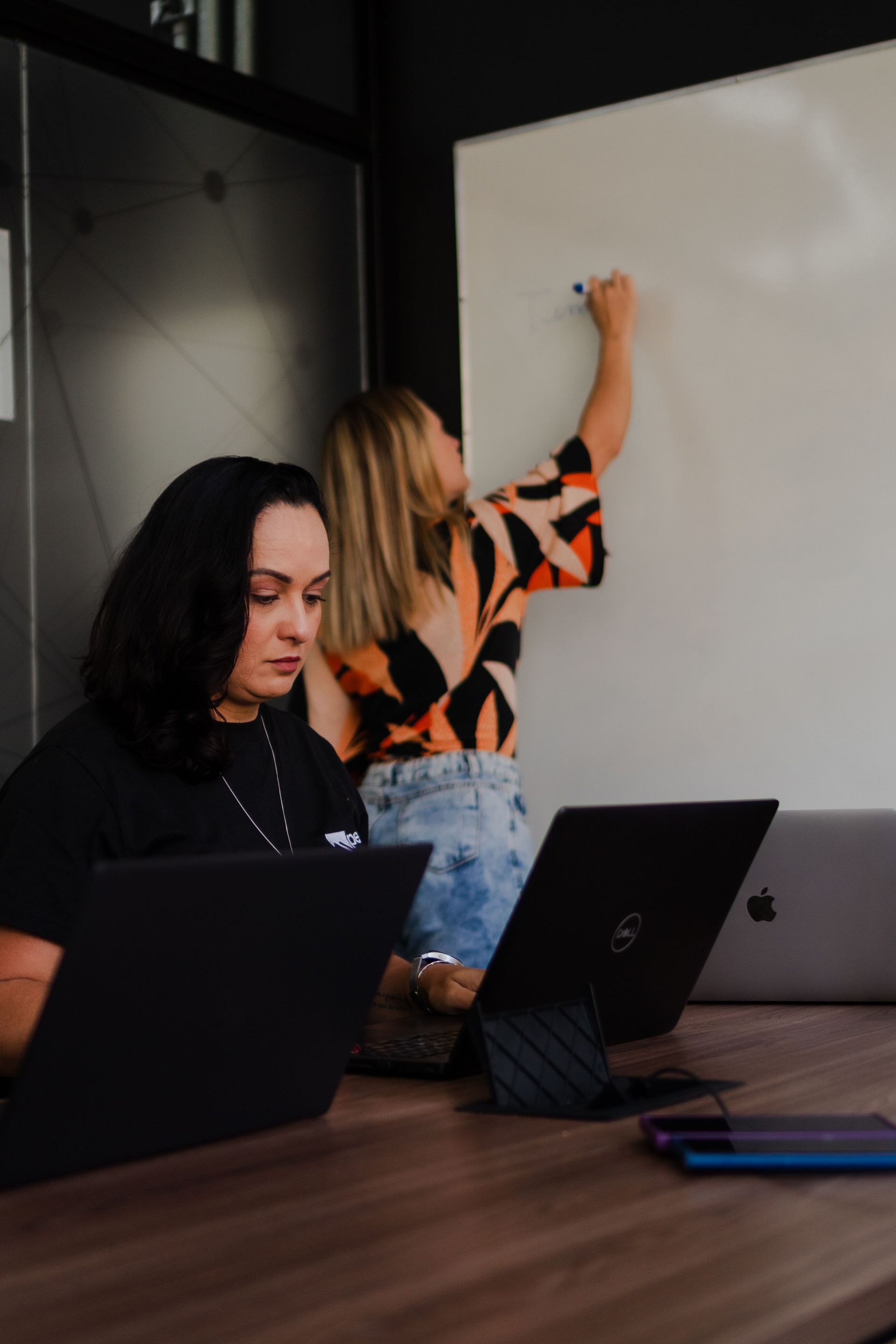 Two people in a meeting room, one writing on a whiteboard and one working on a laptop.