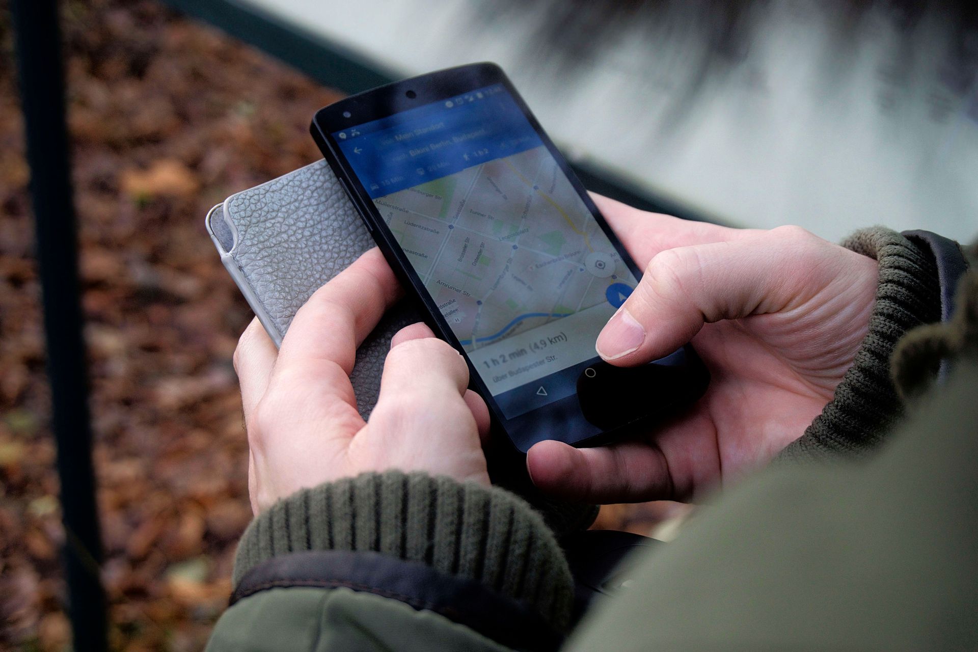 Hands holding a smartphone and a gray wallet outdoors, with a blurred wooded background