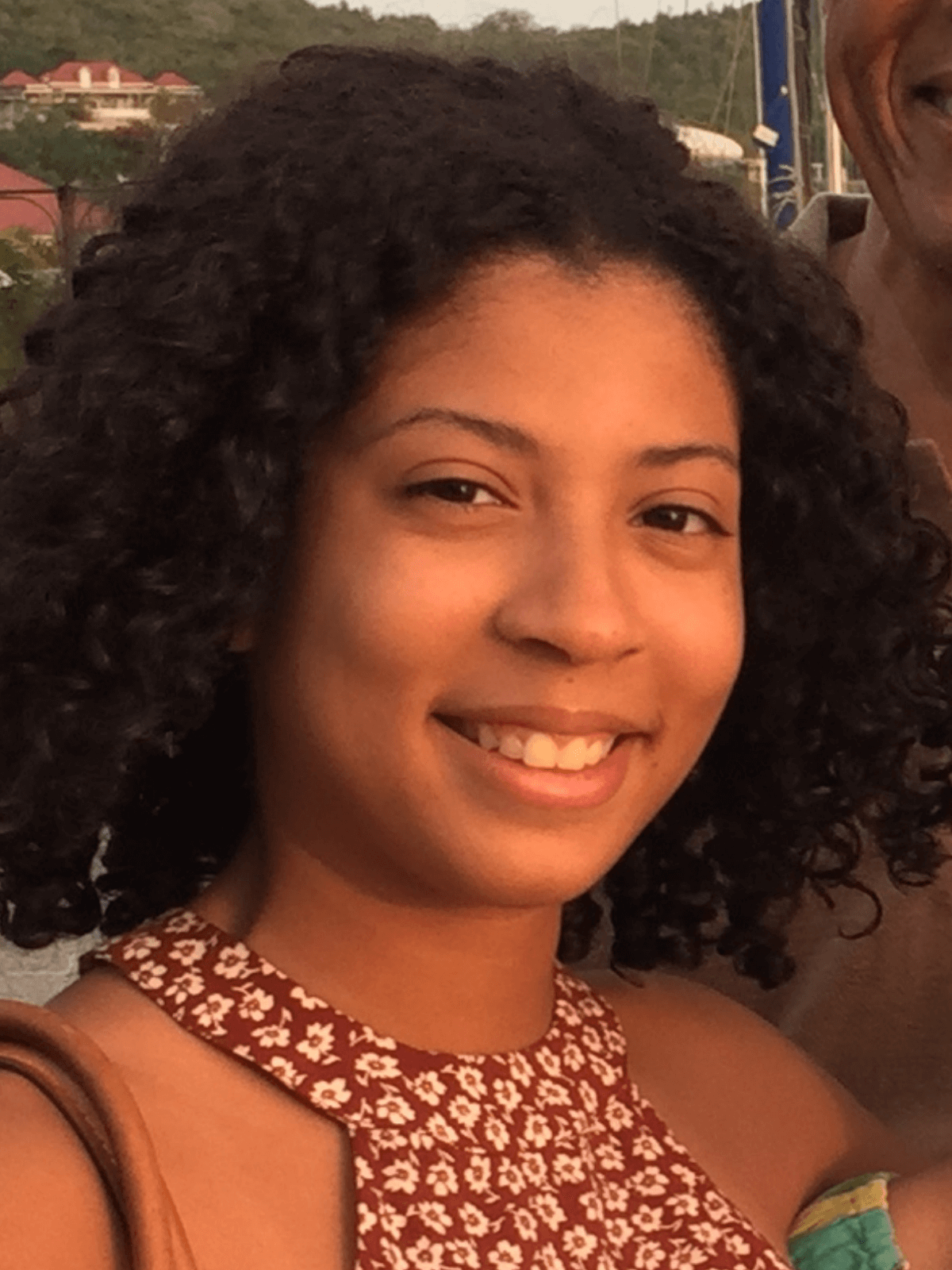 Smiling woman with curly hair outdoors at sunset, wearing a patterned halter top.