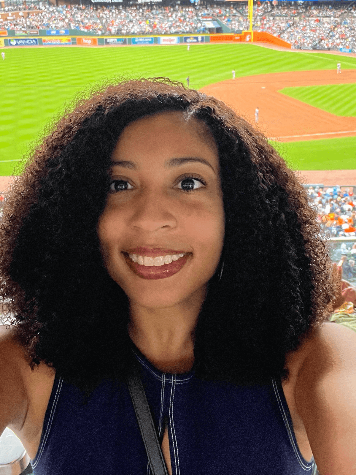 Smiling woman taking a selfie at a baseball stadium with the field and crowd behind her