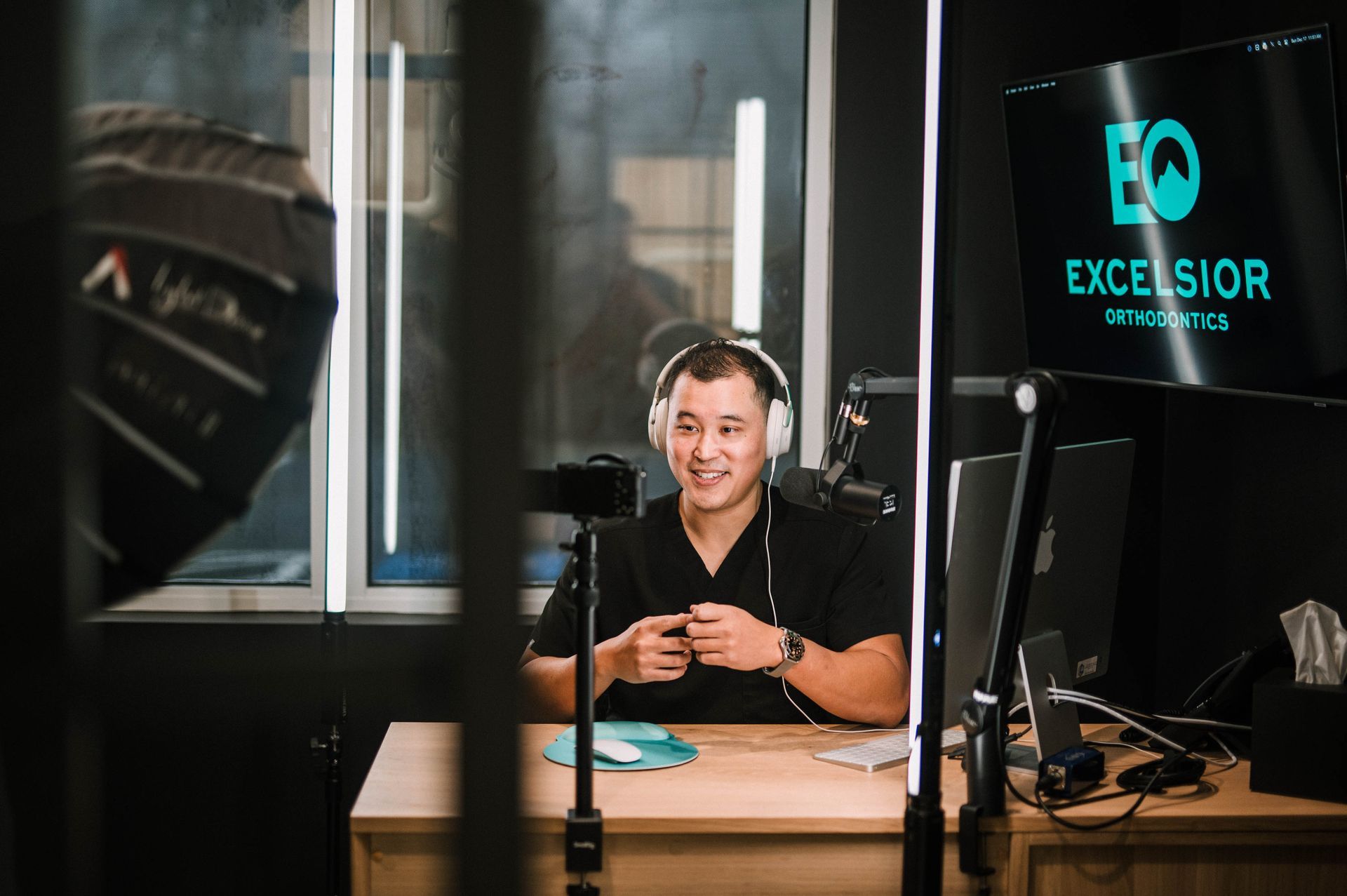 Man at a desk speaking into a microphone in a podcast studio with EXCELSIOR signage.