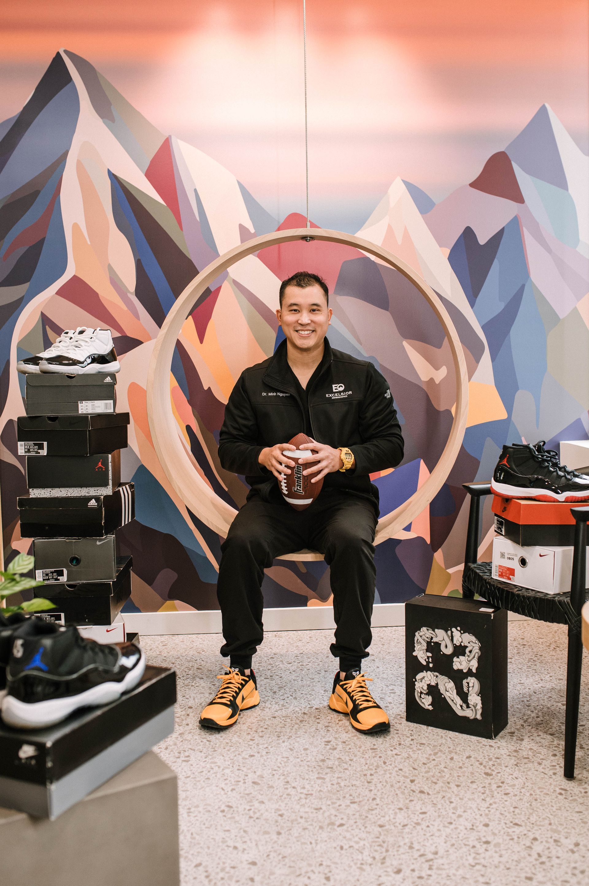 Man seated in a sneaker store, surrounded by stacked shoes, with a colorful mountain mural behind him