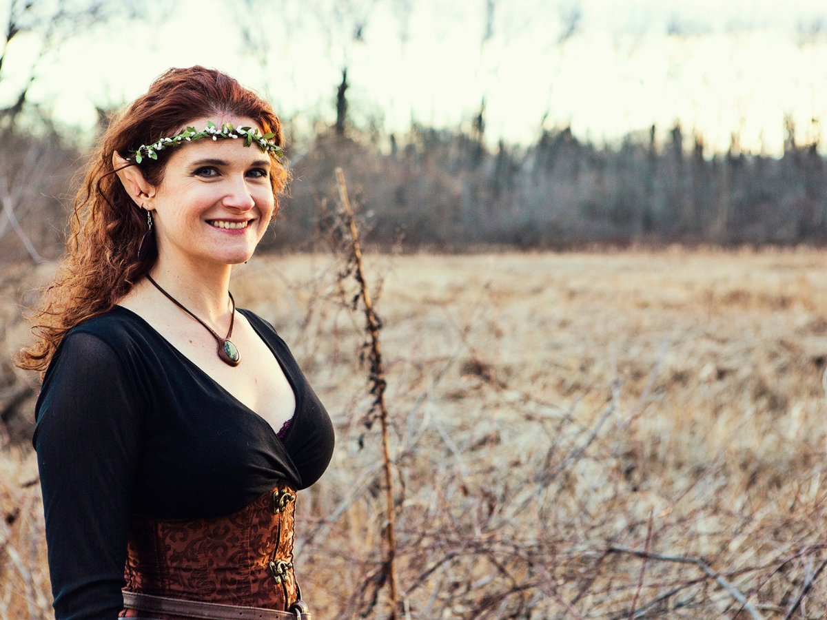 Woman with red hair, wearing a leaf crown and elf ears, smiles in a field.