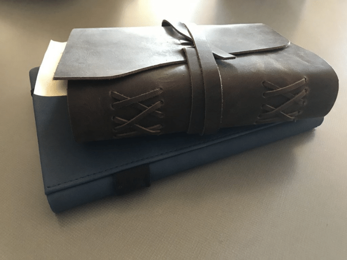 Brown leather-bound journal with strap, resting atop a blue notebook on a table.