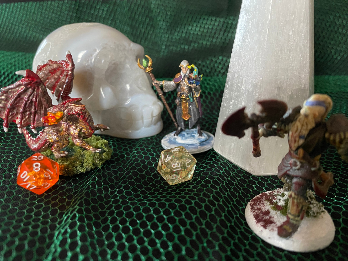 Miniatures and dice on green mesh fabric: skull, wizard, dragon, warrior, crystal, and a d20.