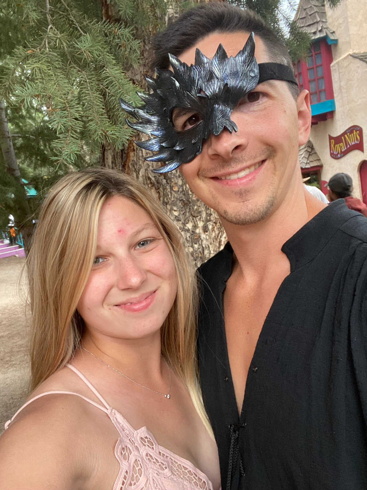 Couple smiling, posing together outdoors. Man wears a spiky mask and black top, woman in a pink lace top.