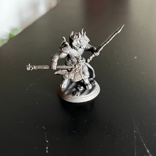 Miniature figure of a creature wielding a sword and a club. It has a crown and is standing on a circular base.