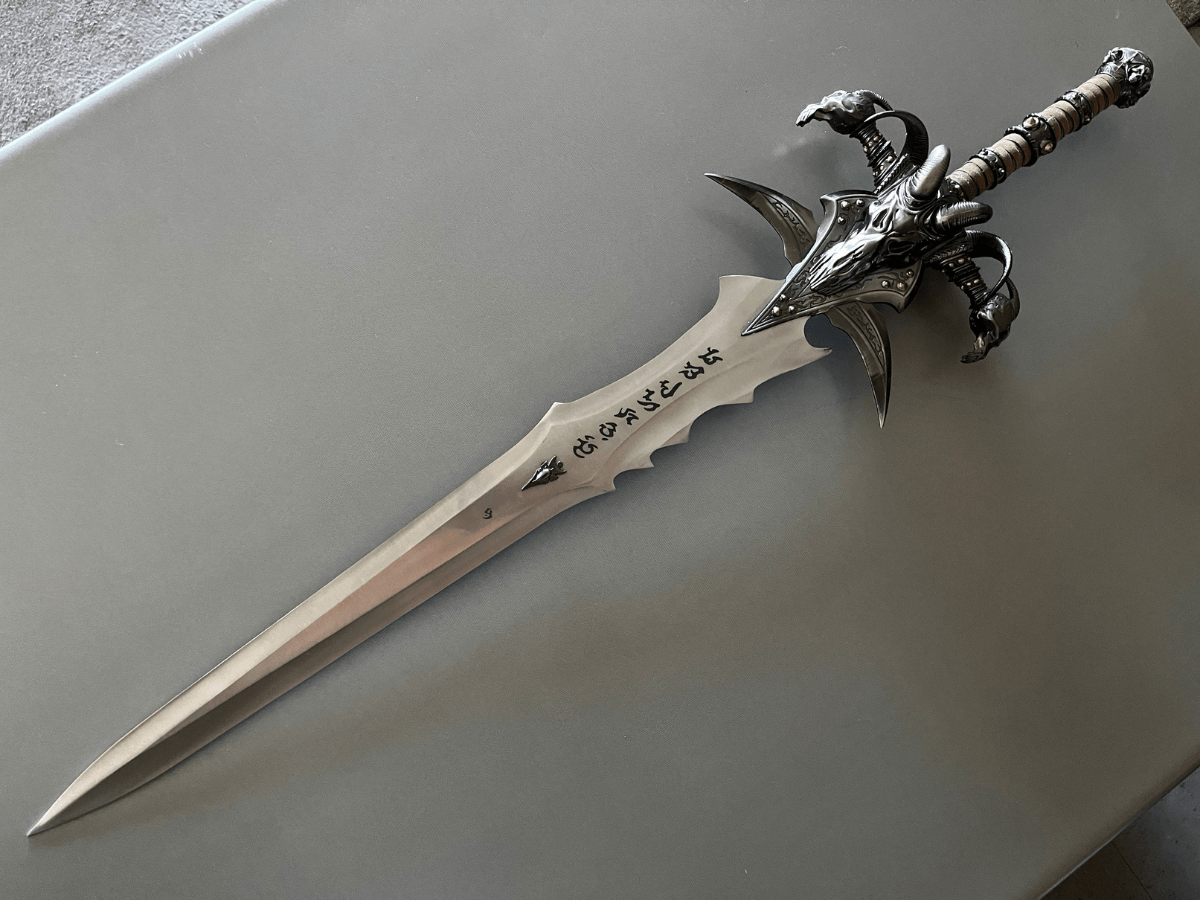 Ornate silver sword with skull-like guard and runic inscription on the blade, resting on a gray surface.