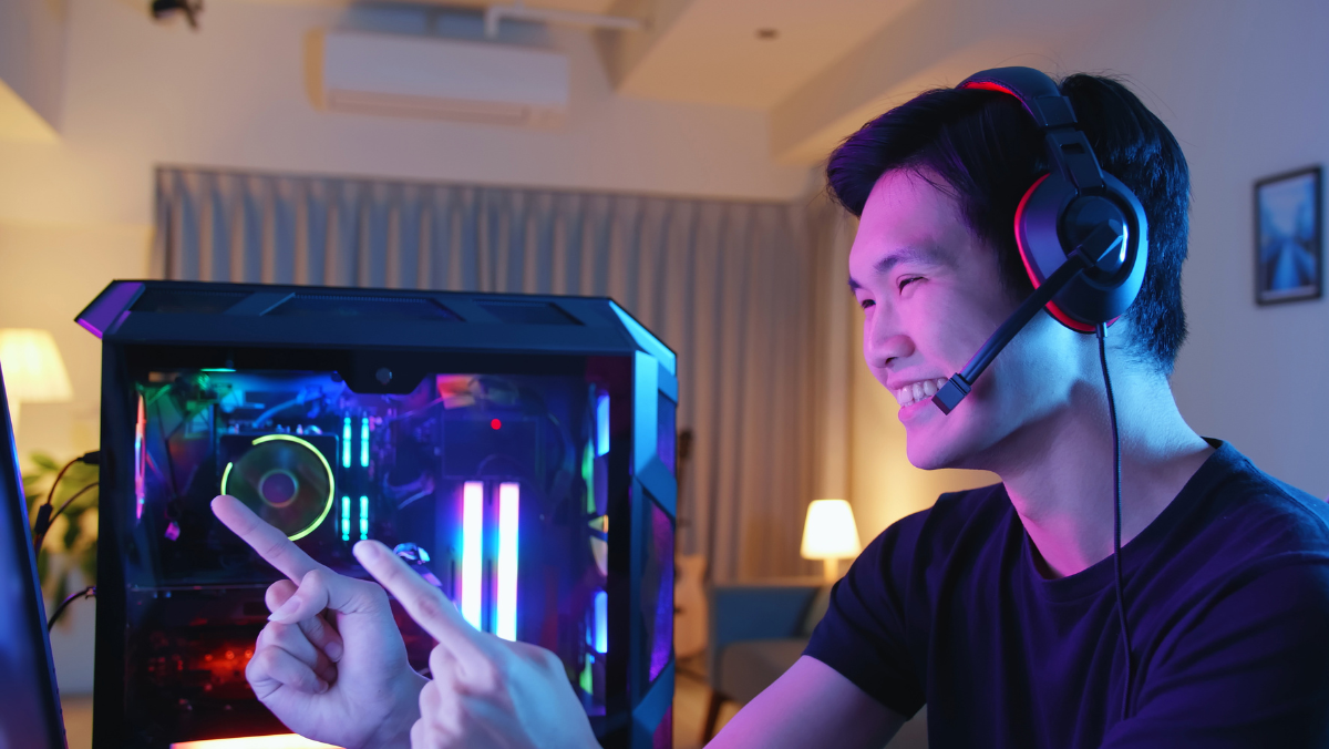 Man with headset smiles, points at glowing gaming computer with colorful internal lights.