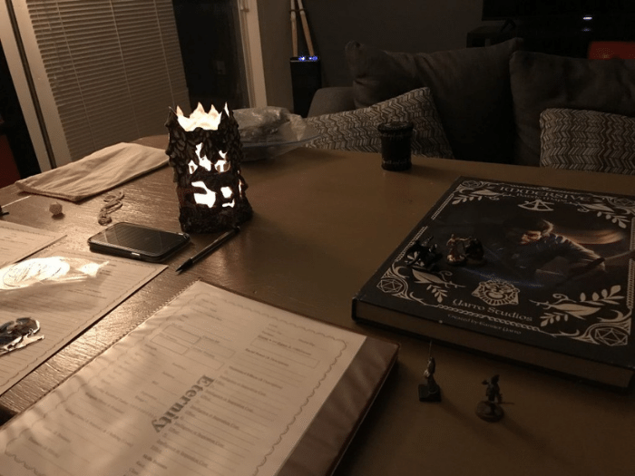 Tabletop RPG setup: open books, dice, miniatures, lit carved candle holder. Dim lighting, indoor setting.