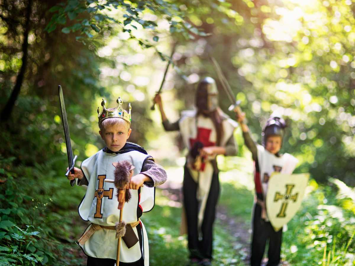 Three children dressed as knights in a forest, holding swords and a shield. One wears a crown.