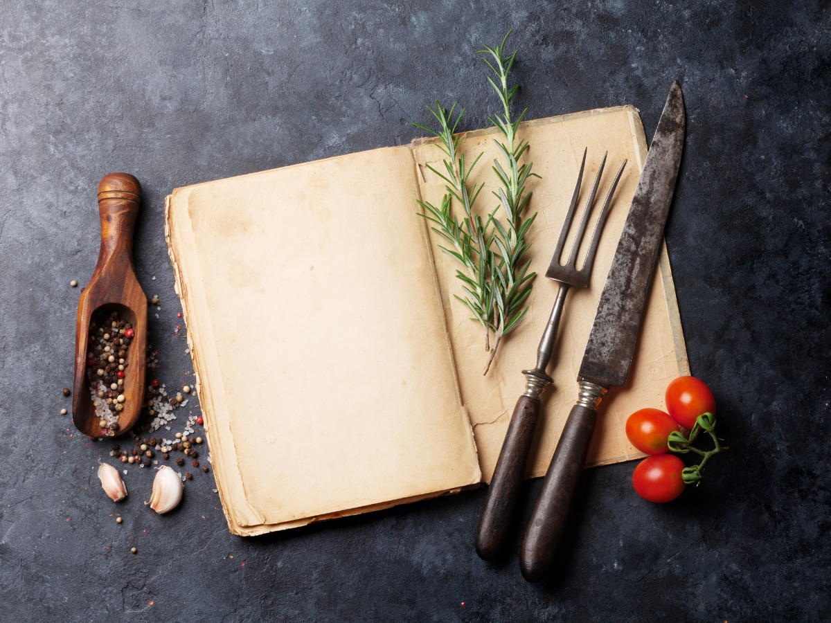 Open cookbook with rosemary, fork, knife, tomatoes, garlic, and spices on a dark surface.
